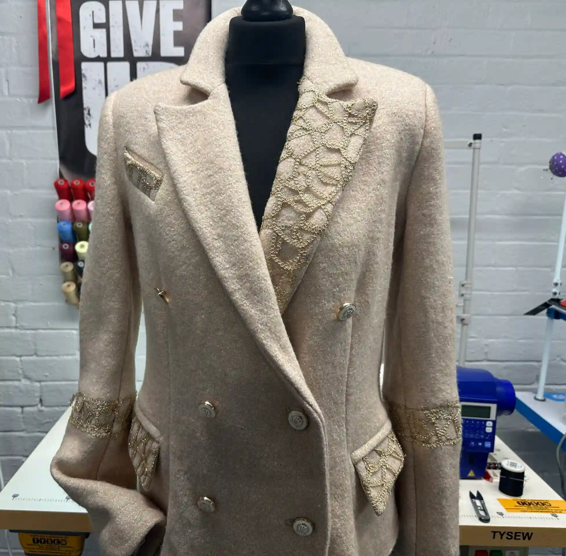A beige textured wool double-breasted K&K Couture coat displayed on a black mannequin, featuring intricate gold metallic lace appliqué on the lapels, pocket flaps, and cuffs, alongside silver buttons, set in a bright sewing atelier with a colourful thread rack and a JUKI sewing machine