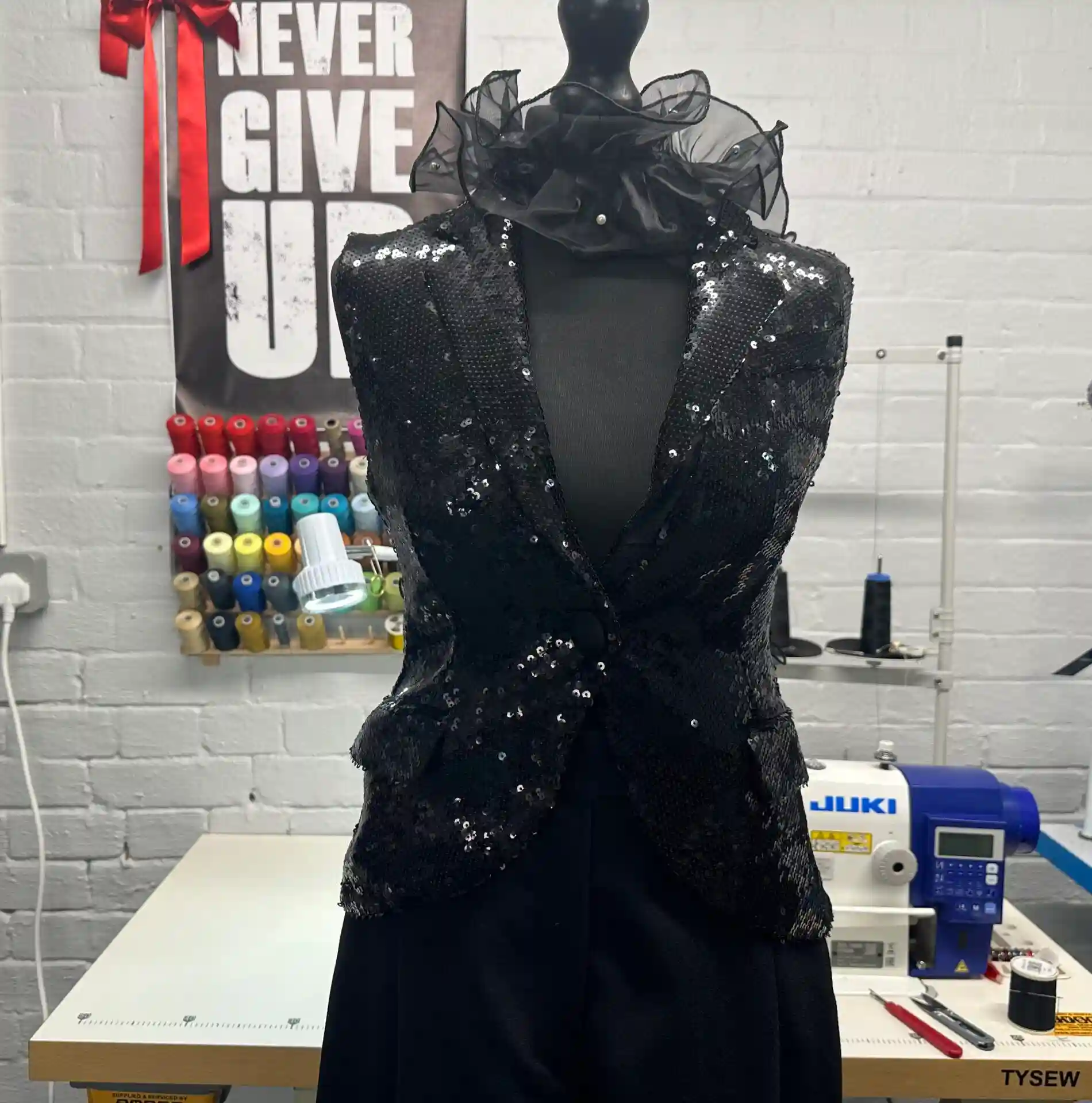 A black mannequin displaying a black sequined K&K Couture vest with a single button and a dramatic sheer organza ruffle collar, set in a bright sewing studio with a colourful thread rack, a JUKI sewing machine, and a "NEVER GIVE UP" poster