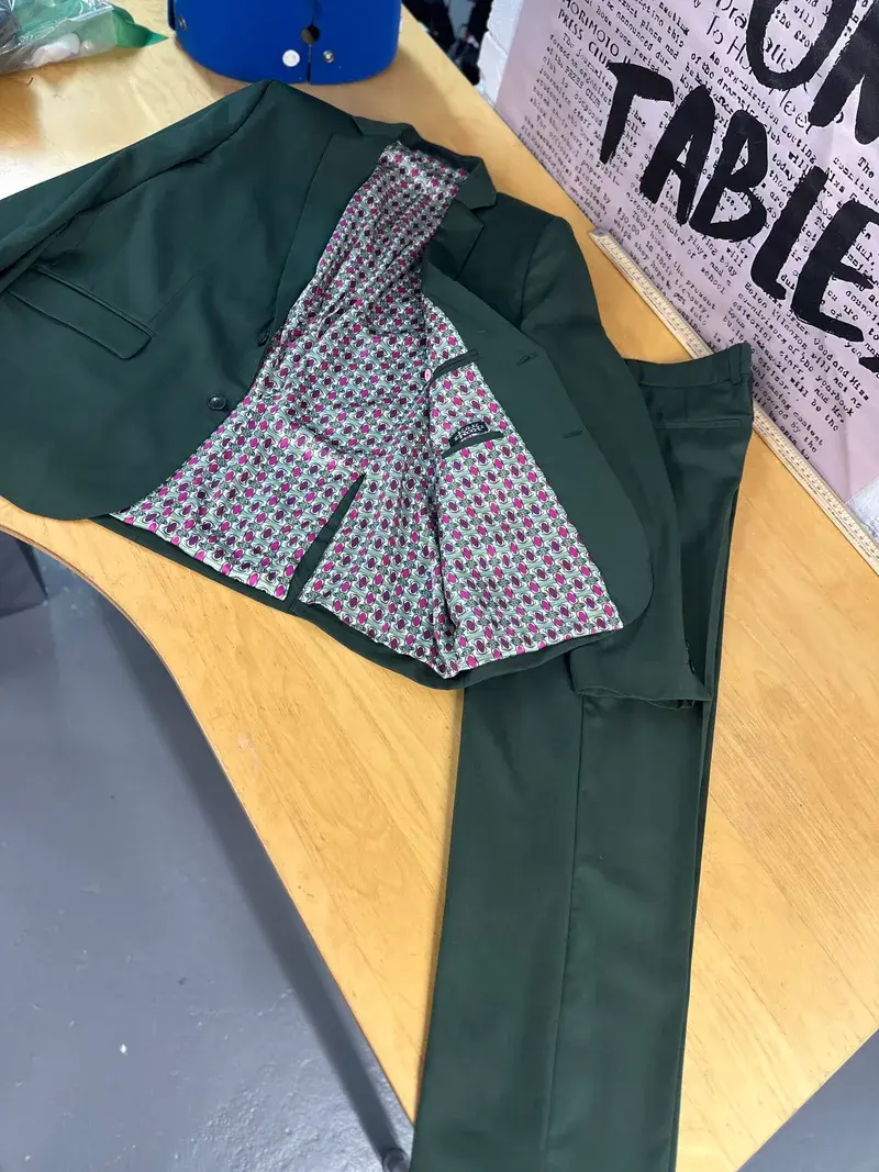 Bespoke men's suit by K&K Couture, featuring a dark forest green jacket and matching trousers laid open on a tailor's workbench, revealing a striking, vibrant custom lining with a pink and grey geometric pattern.