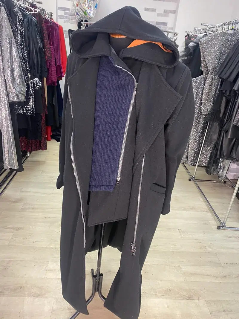 Bespoke menswear and bespoke coat by K&K Couture, featuring a black longline jacket with an attached hood, heavy silver zippers, structured shoulders, and a contrasting blue ribbed knit inner panel, displayed on a mannequin in a boutique