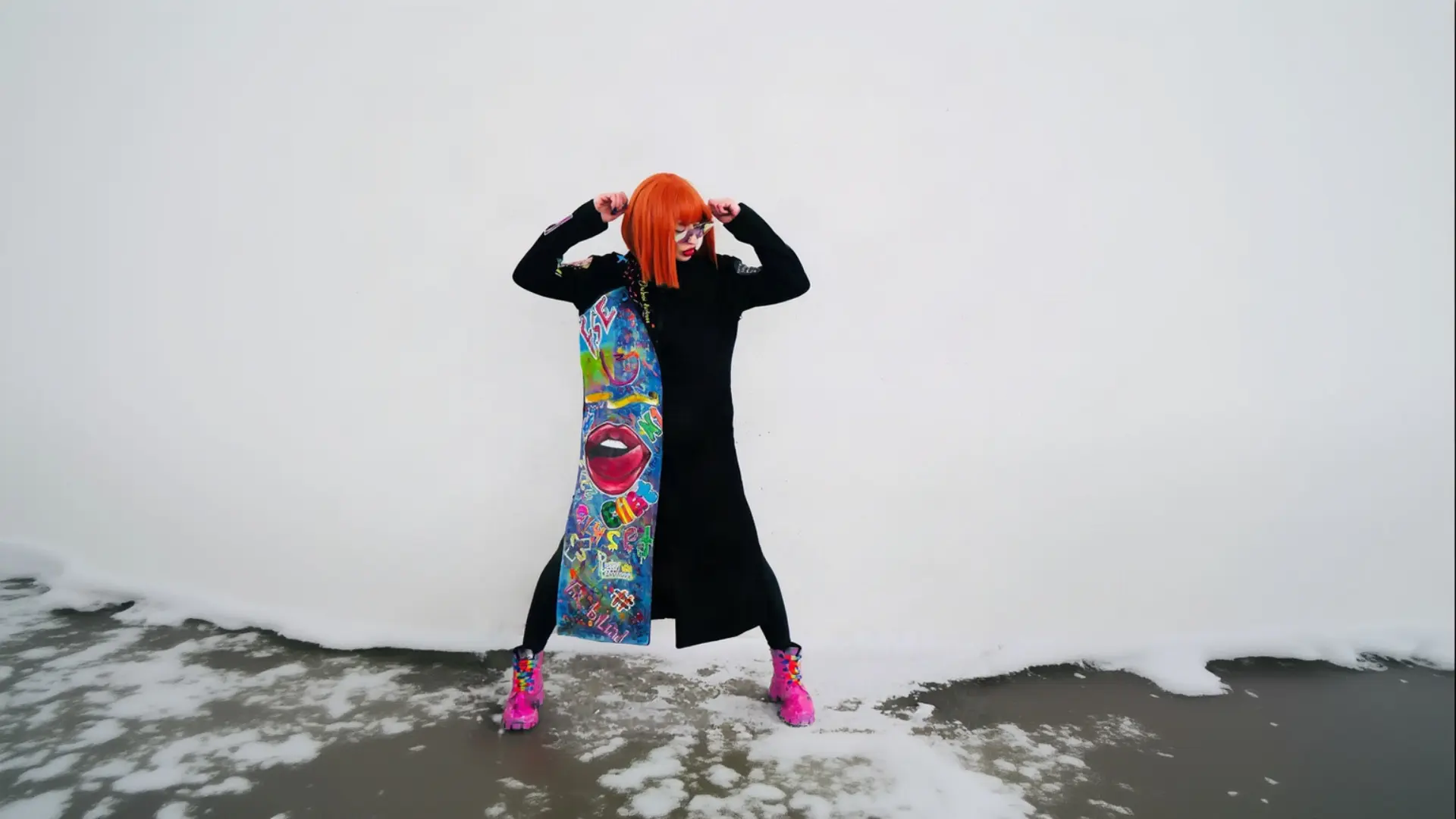 A person with bright orange hair and dark glasses stands with their hands raised near their head, wearing a long black K&K Couture coat-dress featuring a bold, colorful asymmetrical pop-art graphic panel, black leggings, and bright pink patterned boots, against a white wall with patches of snow on the ground