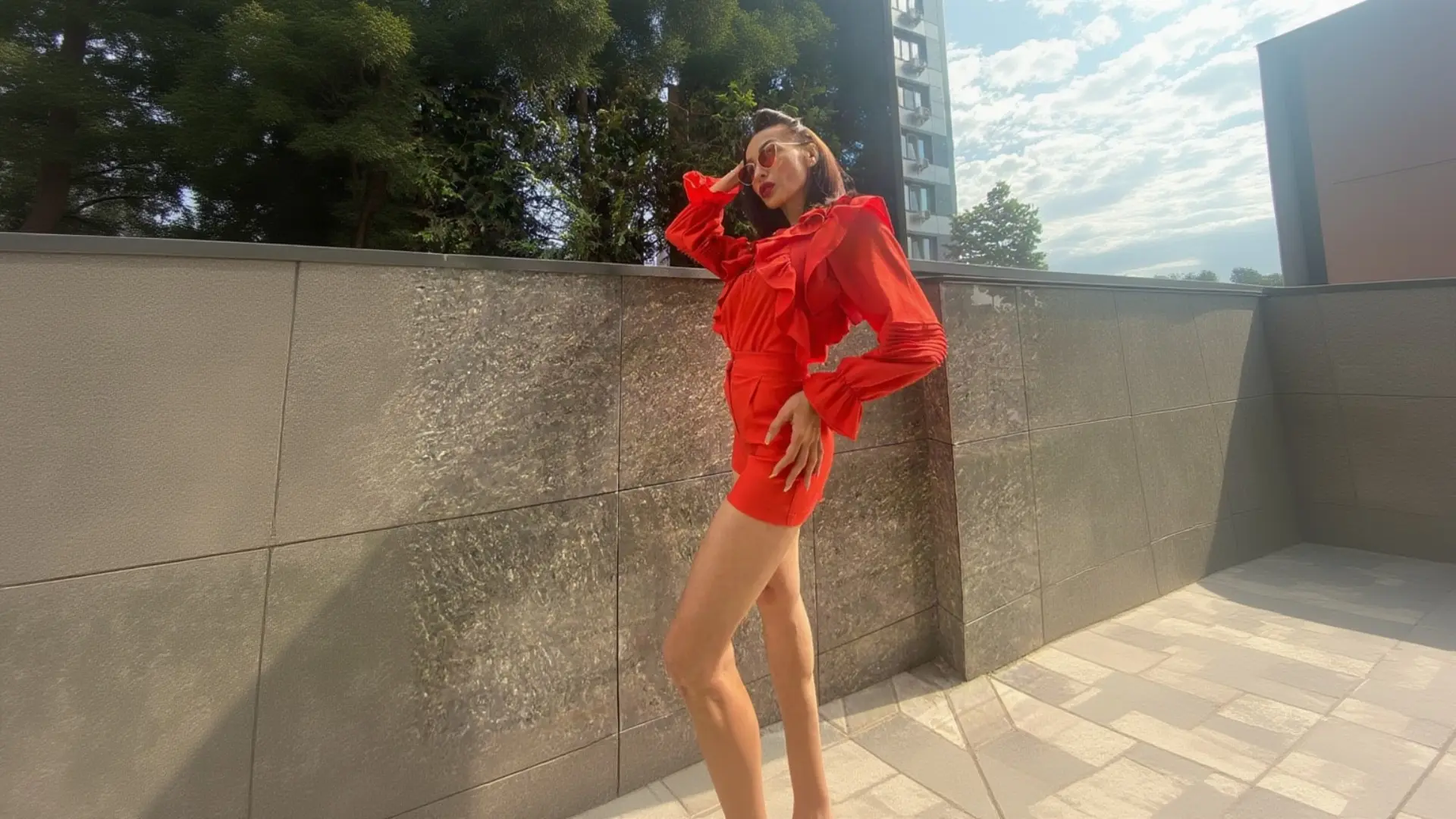 A woman with short dark hair poses confidently outdoors against a grey stone wall, wearing a bright red ruffled long-sleeve top, matching high-slit red shorts, red aviator sunglasses, and red lipstick, with one hand resting on her hip and the other near her face