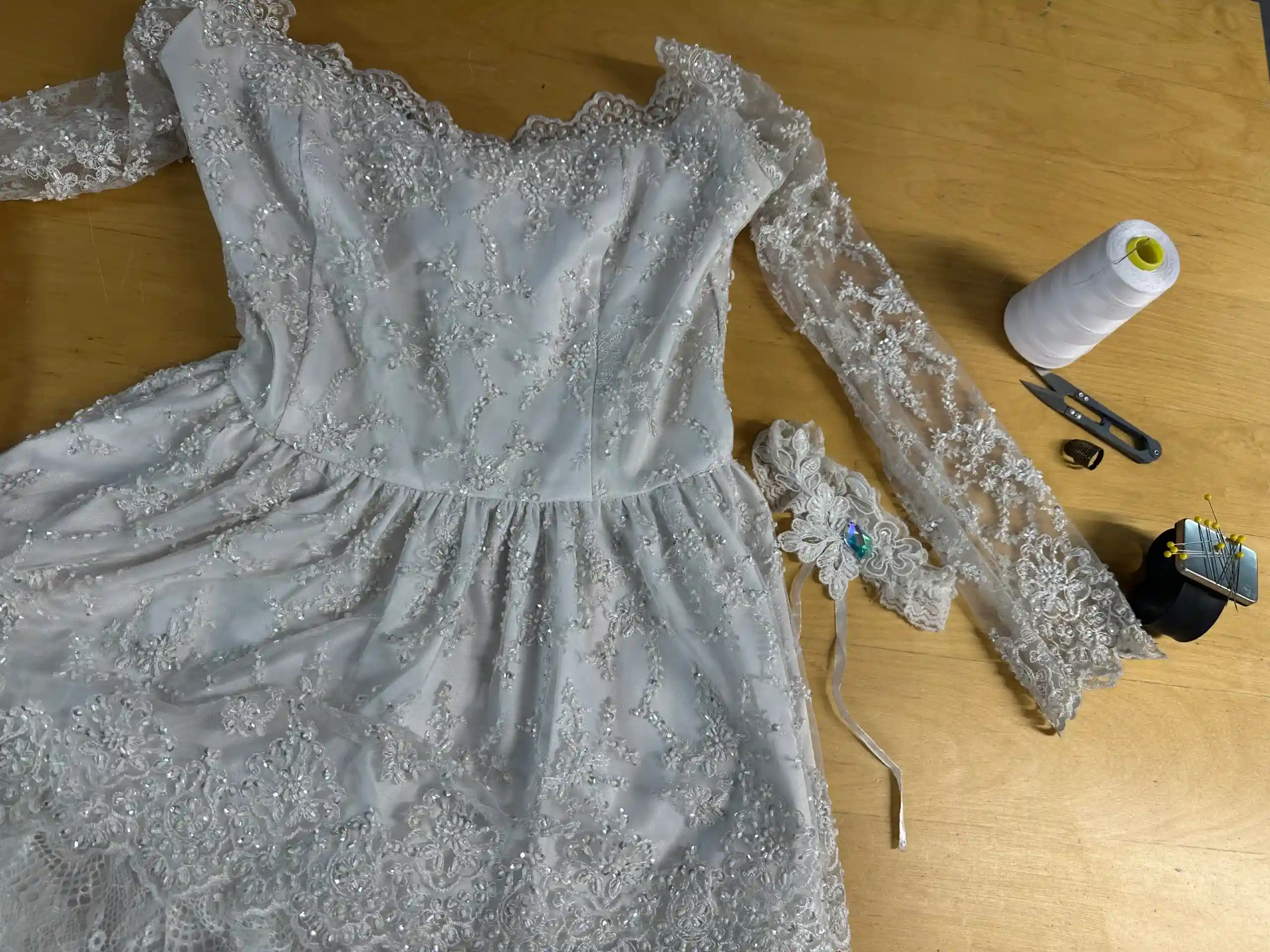 A delicate white lace wedding dress with sheer long sleeves and intricate floral beadwork laid flat on a wooden tailoring table in the K&K Couture atelier. White thread, snipping scissors, a thimble, and a magnetic pin cushion with yellow-head pins rest nearby, alongside a matching lace wrist cuff accessory.