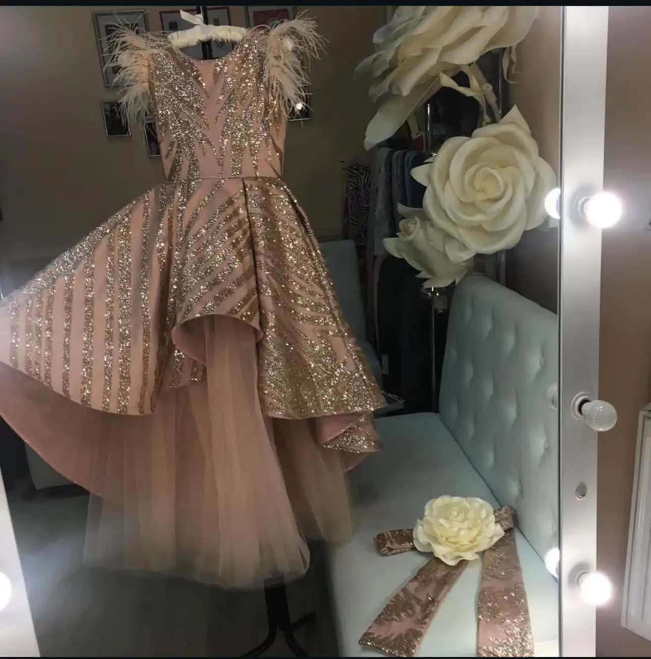 A stunning rose gold sequined gown displayed on a mannequin in a boutique fitting room, featuring feather shoulder accents, a V-neckline, and a dramatic high-low hemline with a tulle underskirt, accompanied by a matching bow with a large white rose resting on a white tufted chair