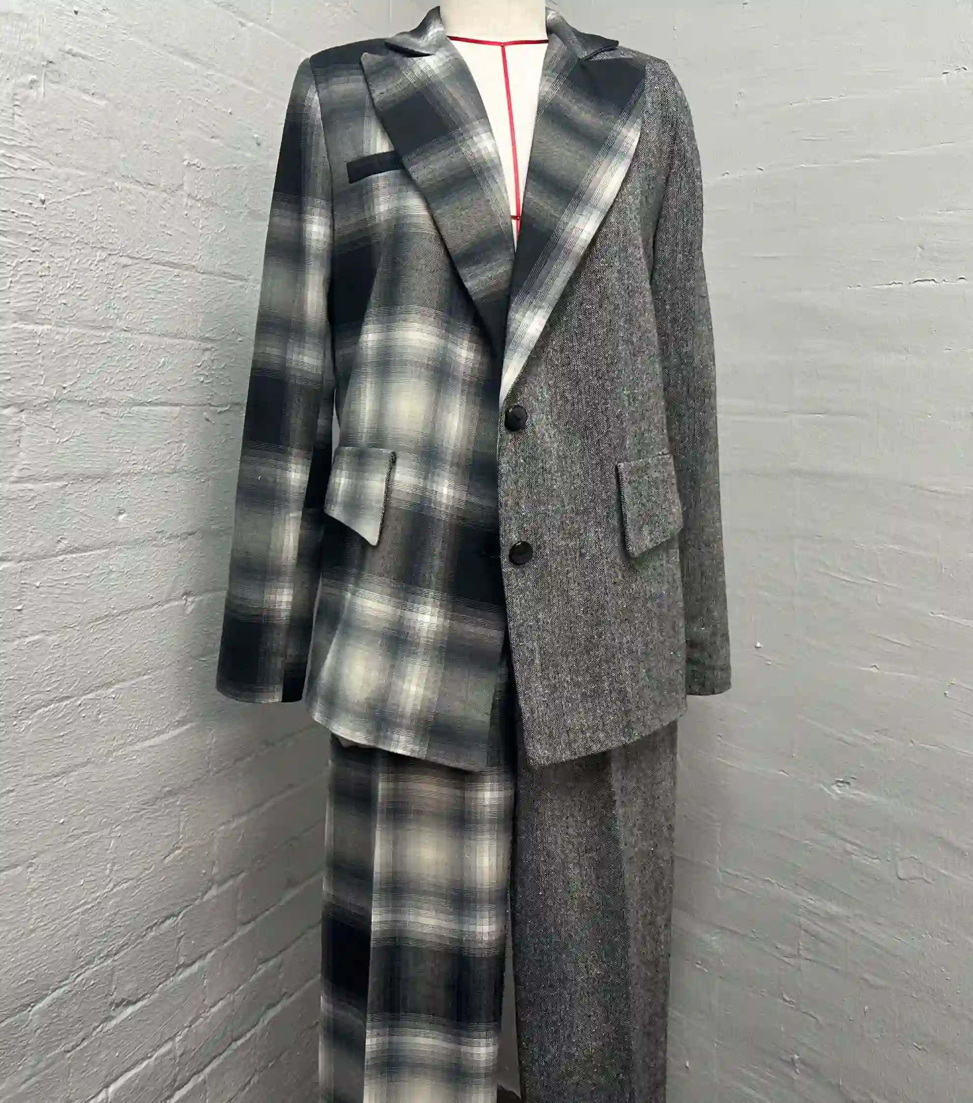 A mannequin displayed against a white painted brick wall wearing a striking K&K Couture suit featuring an asymmetrical split design: one side of the blazer and matching trousers is grey plaid, while the other side is a solid, textured grey wool, accented with black buttons and a thin red neck cord