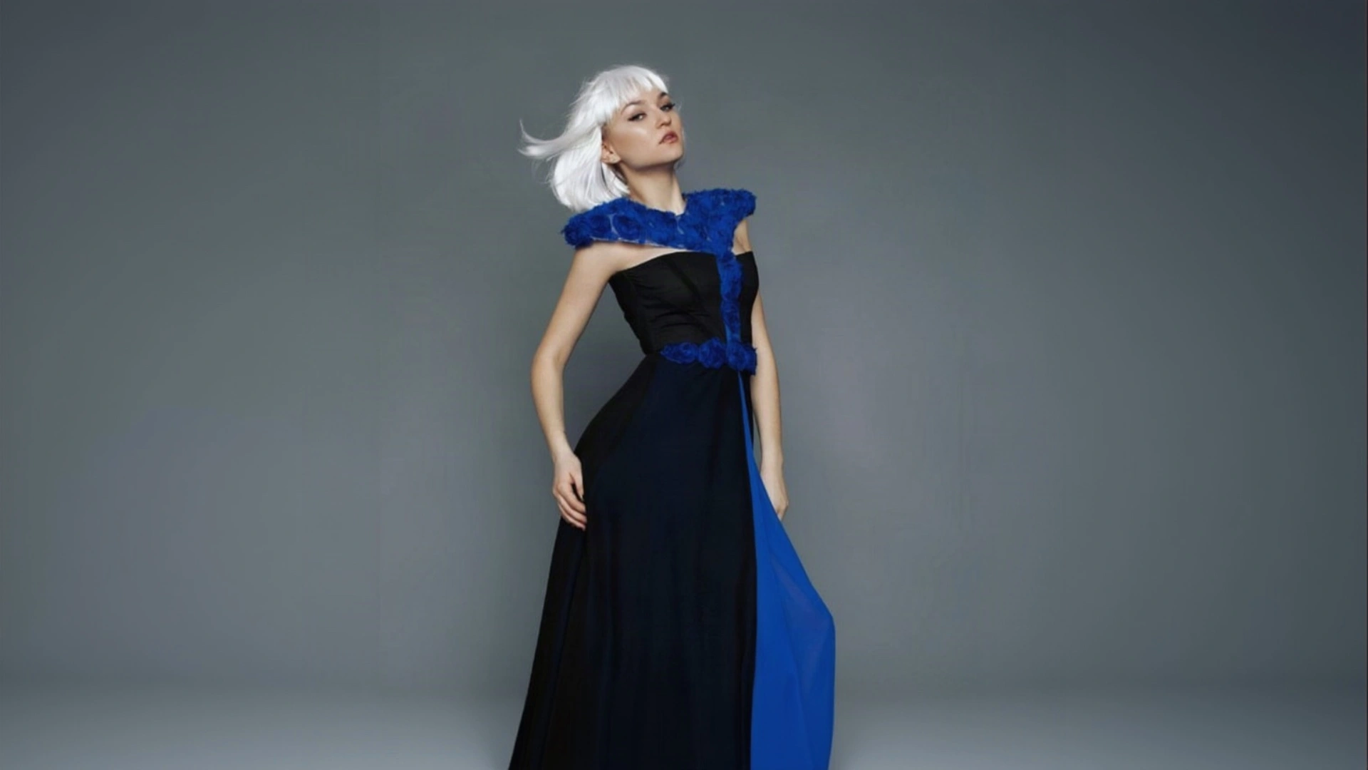 A woman with platinum blonde hair and red lipstick models a striking black and cobalt blue K&K Couture gown featuring a textured blue shoulder wrap, a fitted corset bodice, and a long black skirt with a dramatic blue side panel against a minimalist grey studio backdrop