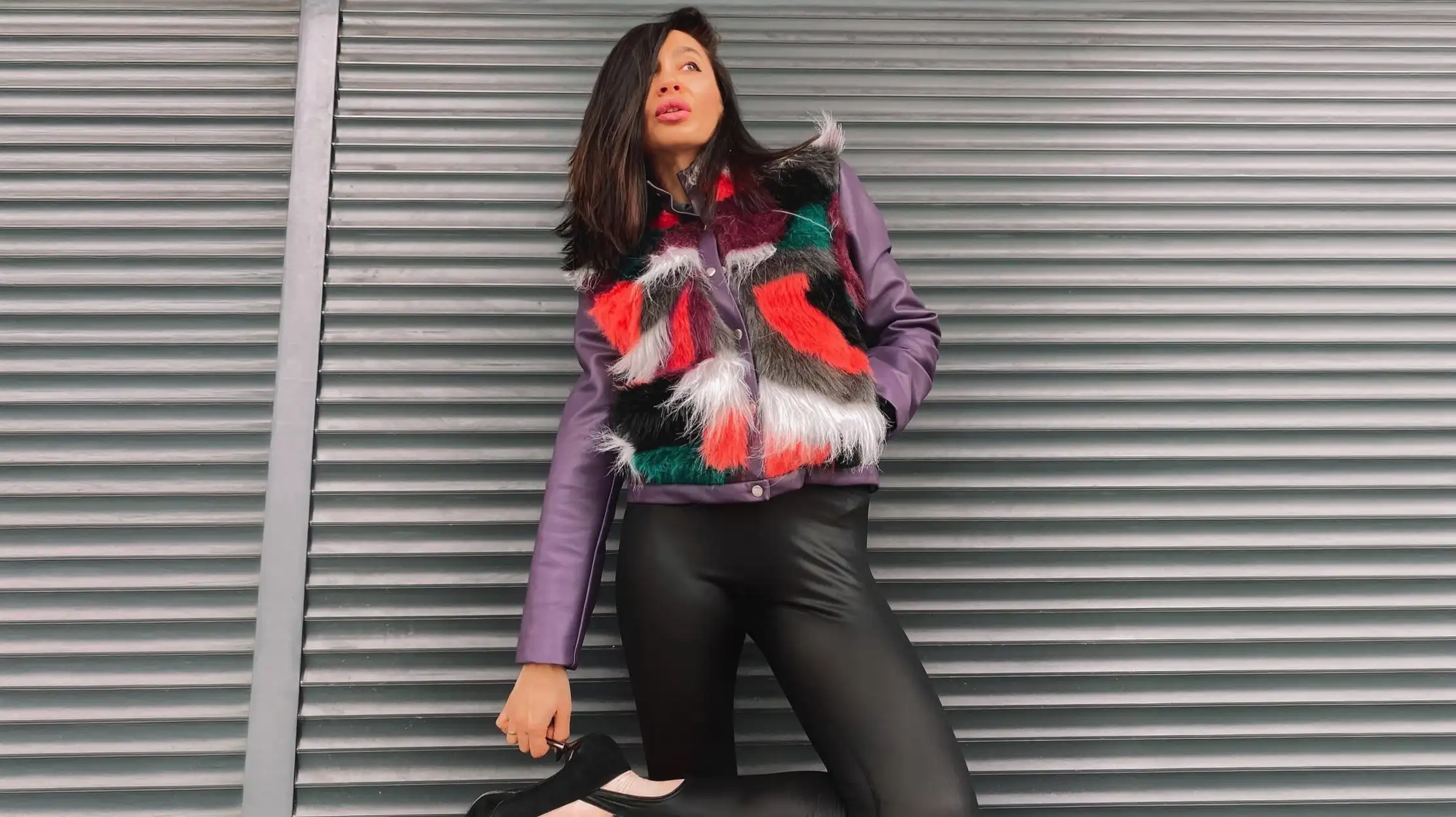 A woman with dark hair and bright pink lipstick leans against a grey corrugated metal wall, holding one black heel in her hand, wearing a purple leather K&K Couture jacket with a vibrant red, green, black, and white shaggy fur body and matching black leather leggings