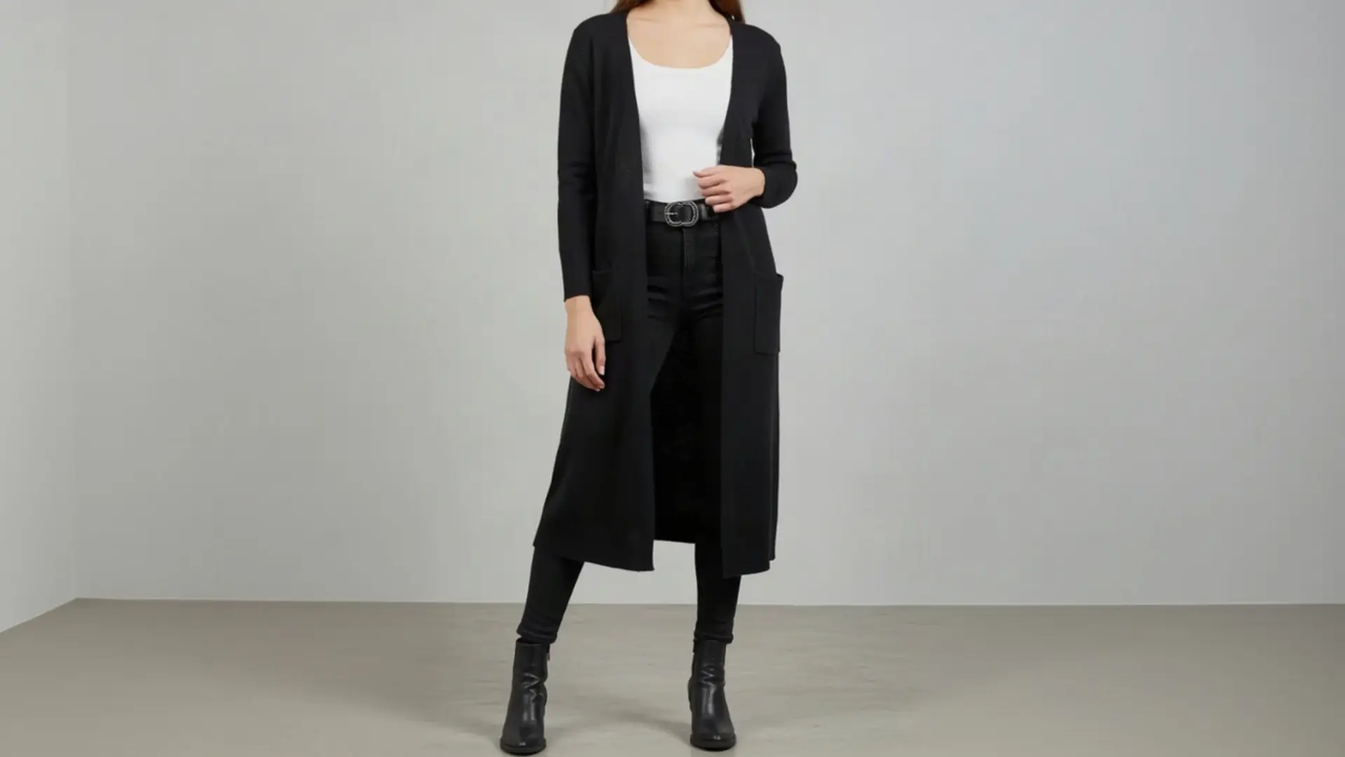 A woman wearing a long black K&K Couture open-front cardigan over a white scoop-neck top, black skinny jeans, a black belt with a silver buckle, and black ankle boots, standing against a minimalist grey studio background