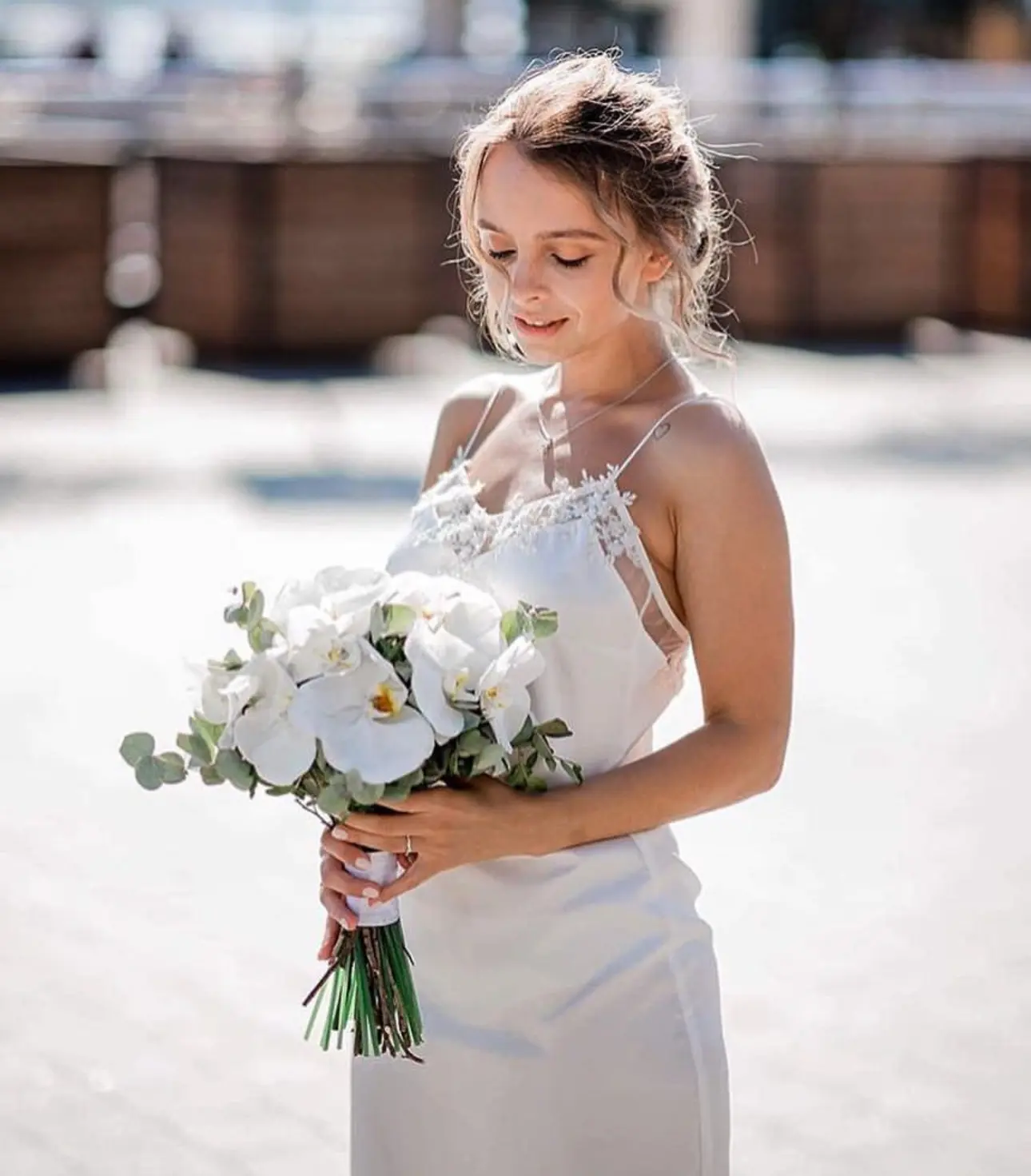 A young woman with her hair in a soft, messy updo stands in bright sunlight, looking down with a gentle smile at a bouquet of white orchids. She is wearing a delicate white K&K Couture slip wedding dress with thin spaghetti straps and intricate lace detailing along the neckline