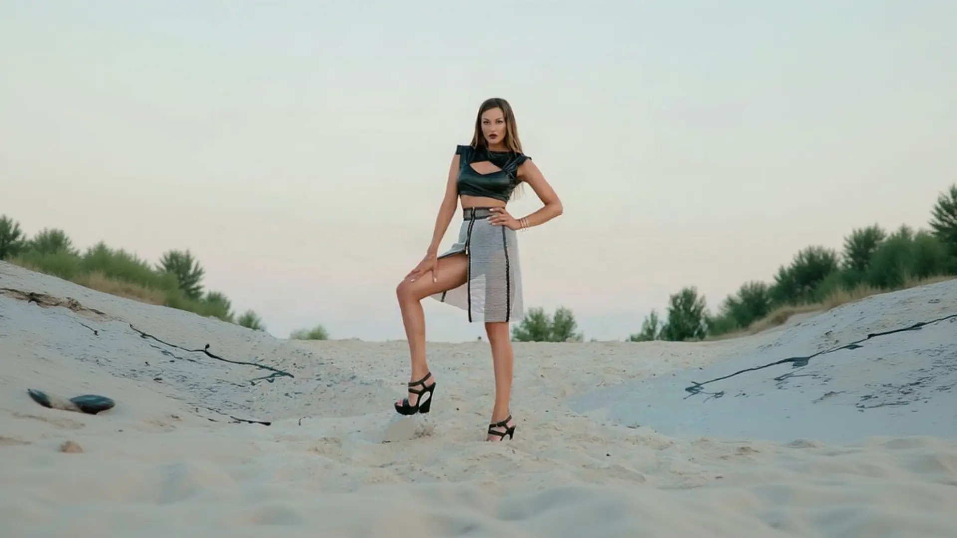 A woman with long hair and bold dark lipstick strikes a confident pose on a sandy landscape at dusk, wearing a black leather-look crop top with a cutout, a high-waisted black and white checkered skirt with a dramatic slit, and strappy black platform heels, showcasing a stunning outfit from K&K Couture