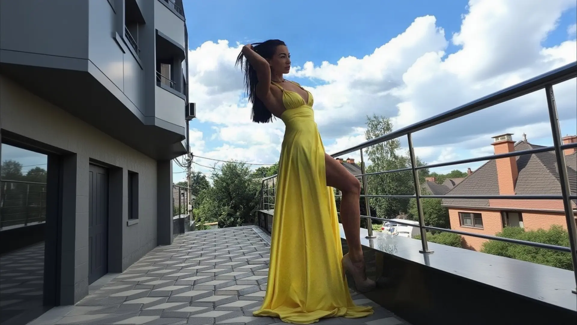 A woman with long dark hair poses gracefully on a modern balcony, lifting her hair with one hand and resting her leg on the ledge, wearing a vibrant yellow K&K Couture halter maxi dress with a plunging neckline and dramatic thigh-high slit, paired with nude platform heels under a bright blue sky with fluffy clouds