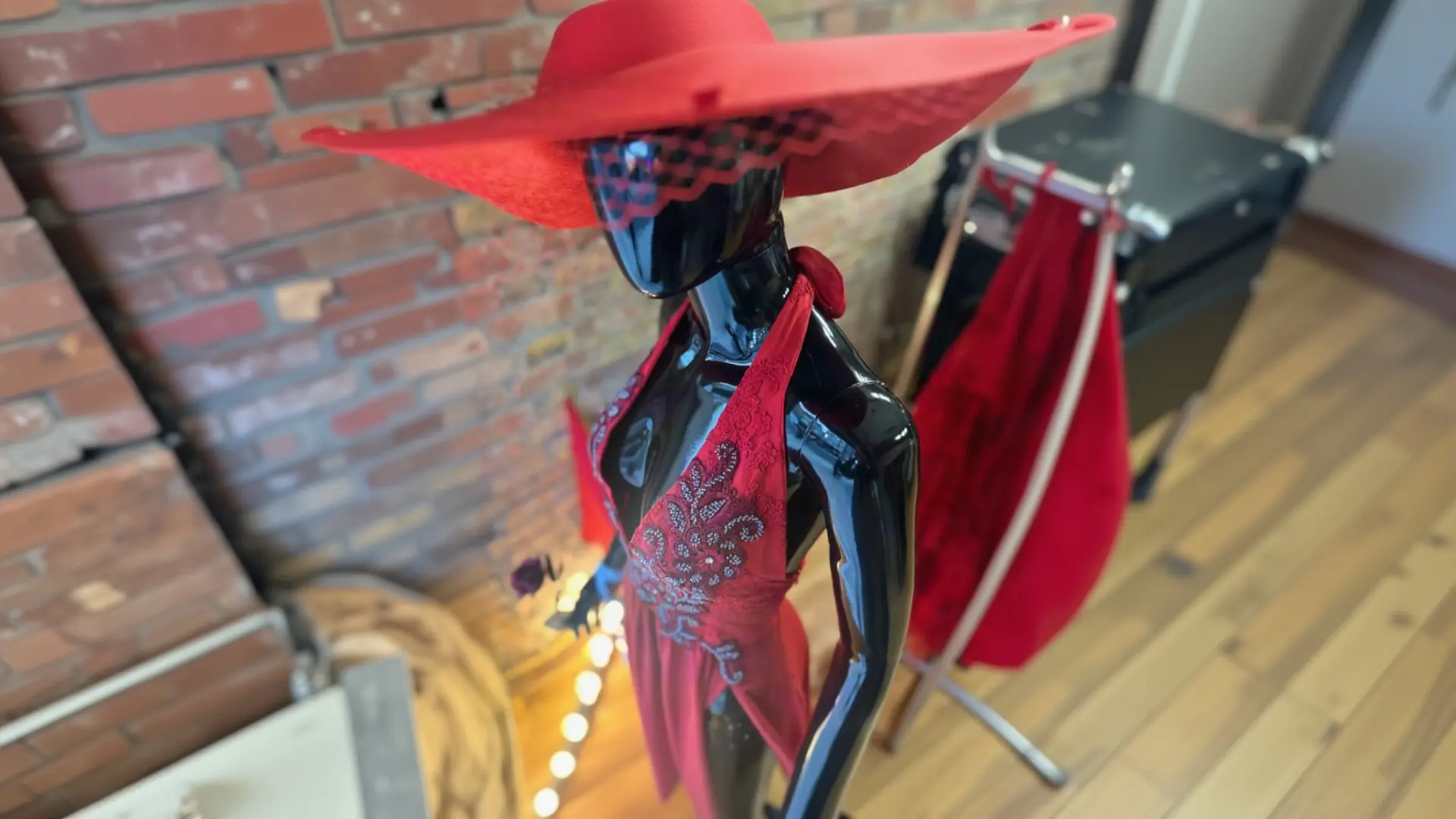 A glossy black mannequin displaying a bold, wide-brimmed red hat with a checkered band and an embellished red halter dress, set against a red brick wall and a red fabric rack inside a boutique featuring K&K Couture pieces