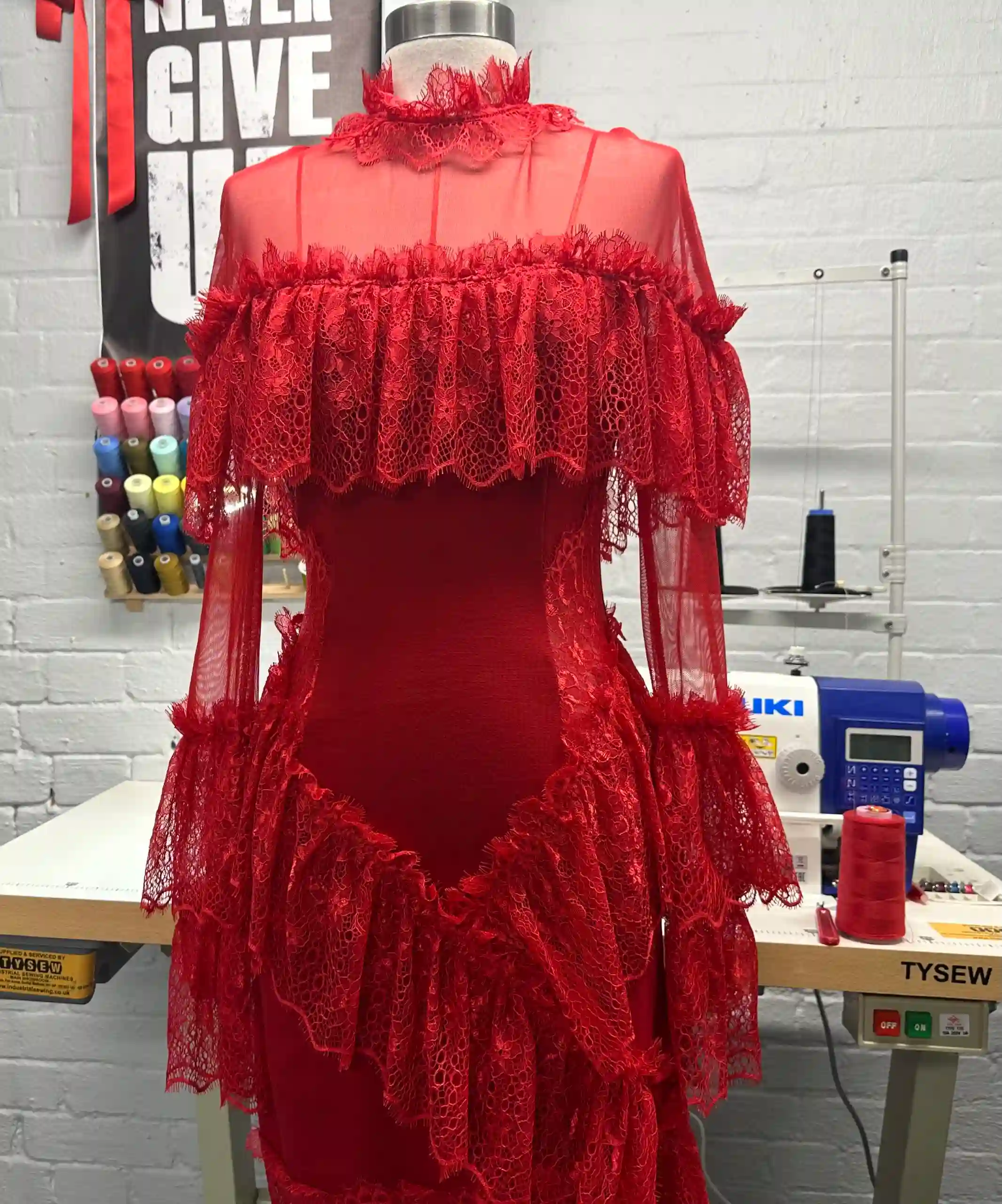 A striking red K&K Couture sheer lace ensemble displayed on a white dress form in a design studio. The garment features a high lace collar, a dramatic off-the-shoulder lace ruffle, long sheer sleeves with ruffled cuffs, a fitted sheer bodice over a solid red base, and a voluminous tiered lace peplum overlay. It is set against a white painted brick wall with a colorful thread rack, a JUKI sewing machine, and a "NEVER GIVE UP" poster in the background