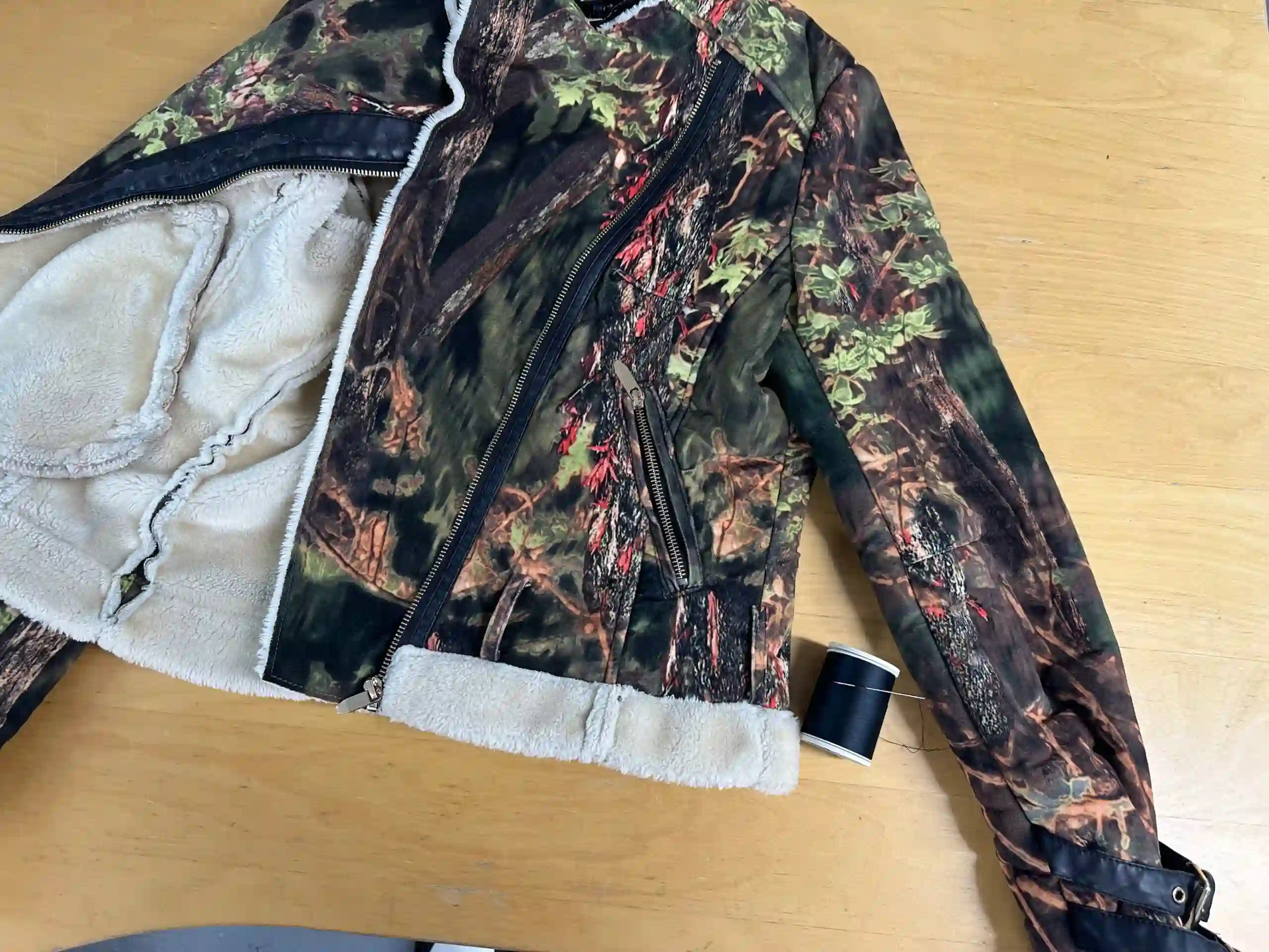 A camouflage print jacket featuring a thick, plush white sheepskin lining and black leather trim lays flat on a wooden workbench in the K&K Couture atelier. The heavy-duty jacket features black zippers, and a spool of black thread rests nearby, indicating an alteration in progress.