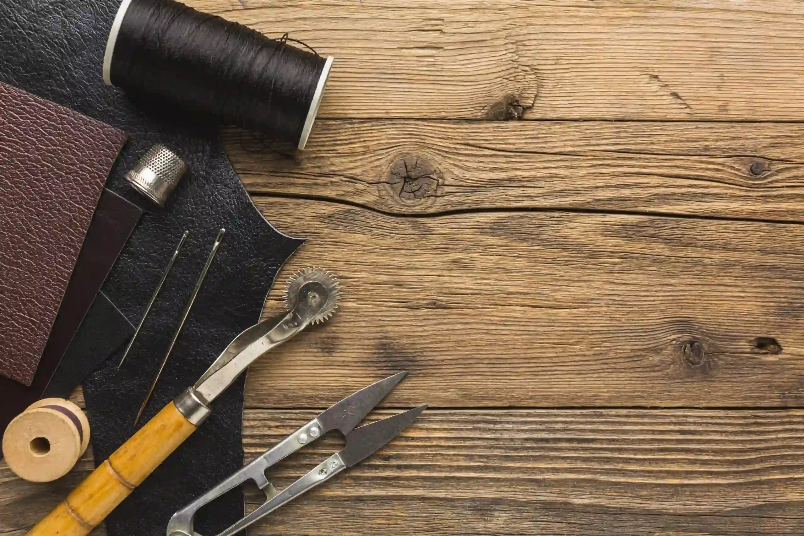 A flat lay of specialized leather alteration tools on a rustic wooden workbench at the K&K Couture atelier, featuring black and burgundy leather swatches, a large spool of heavy-duty black thread, a metal thimble, a wooden-handled tracing wheel, and heavy-duty tailoring snips.