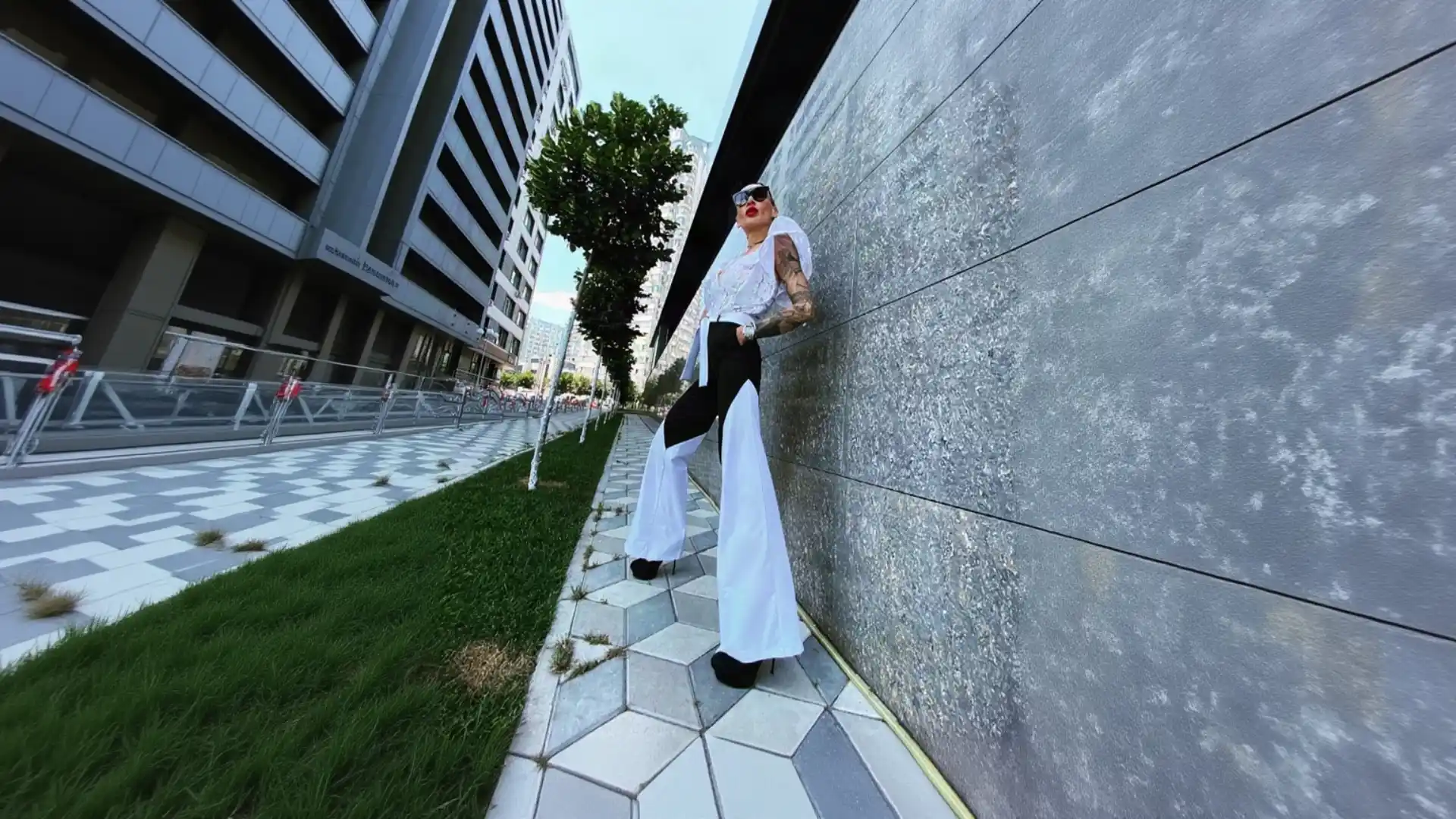 A woman with long dark hair, tattoos, and red lipstick leans confidently against a textured grey stone wall in a modern city. She is wearing custom K&K Couture bespoke trousers featuring a bold color-block design with a fitted black upper section and dramatic, voluminous white flared hems, paired with a white ruffled top, sunglasses, and black platform heels