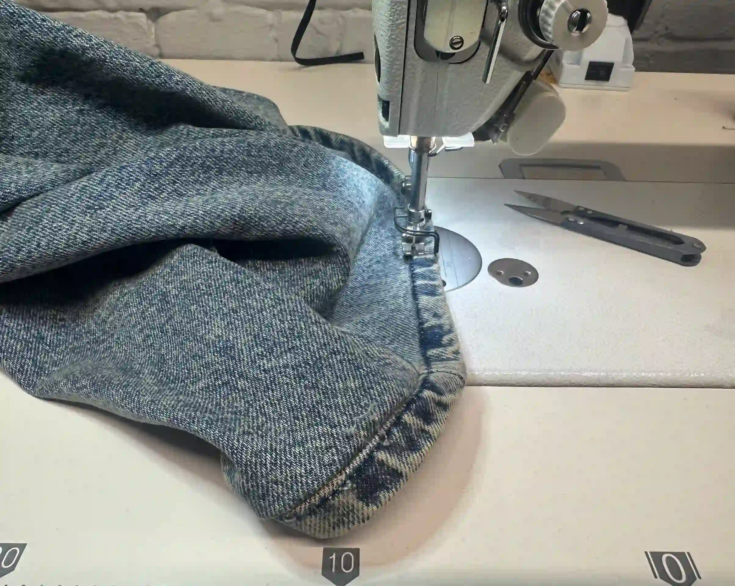 A close-up view of a heavy-duty industrial sewing machine stitching the hem of blue denim jeans in the K&K Couture atelier. The thick, bunched denim fabric is positioned under the needle, with a pair of thread snips resting on the white worktable against a white-painted brick wall.