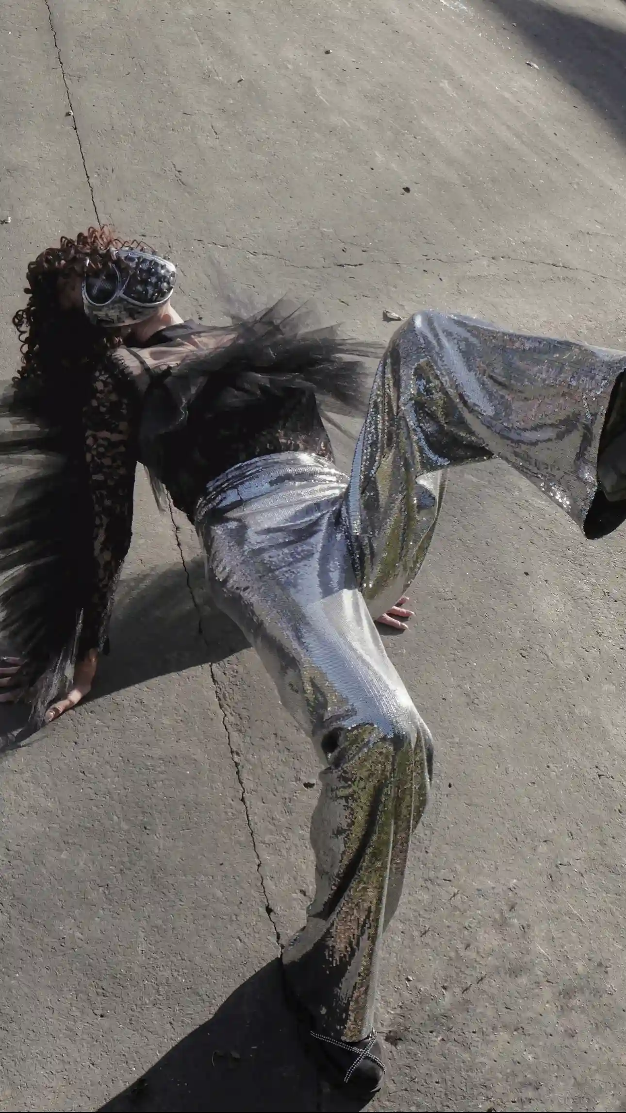 A dramatic, high-fashion portrait of a woman lying on a concrete surface, wearing a studded mask, a black top with voluminous tulle sleeves, and shimmering silver sequin wide-leg trousers from K&K Couture. The pose highlights the dramatic flare and metallic sheen of the custom-fitted trousers.