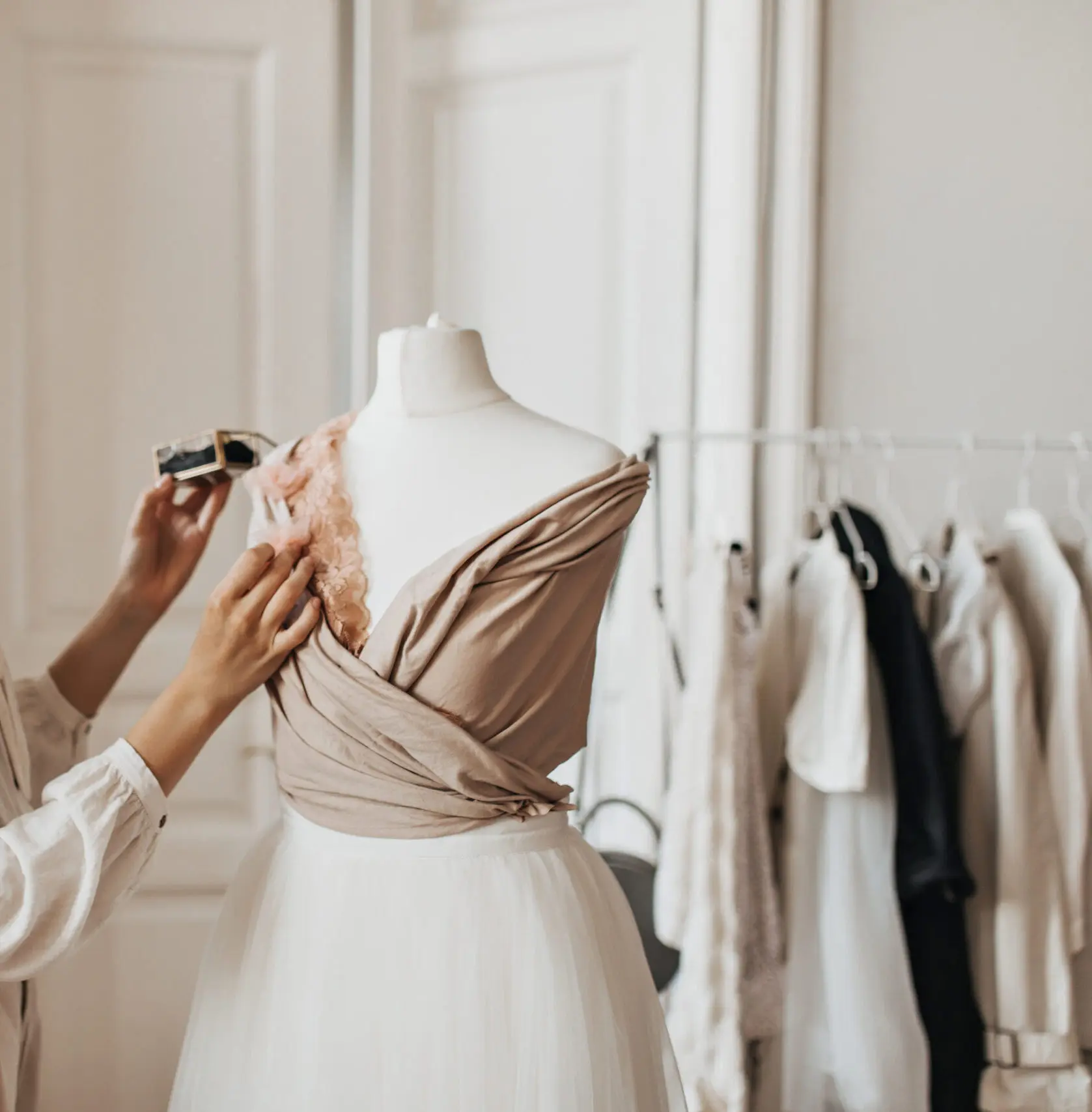 A close-up of a tailor's hands carefully adjusting a delicate pink lace appliqué and a beautifully draped beige bodice on a white dress form with a white tulle skirt. The scene is set in a bright, elegant K&K Couture atelier with a clothing rack featuring neutral-toned garments softly blurred in the background