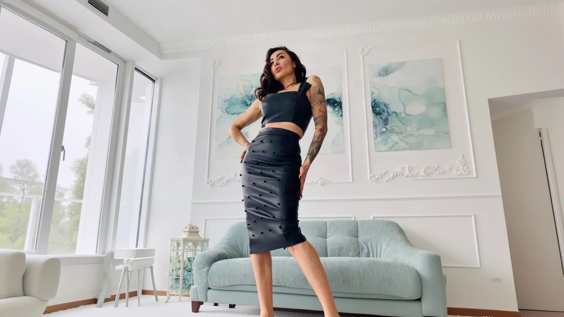 A woman with dark hair and arm tattoos poses confidently with her hands on her hips in a bright, elegant living room, wearing a black K&K Couture matching set featuring a square-neck crop top and a studded midi pencil skirt