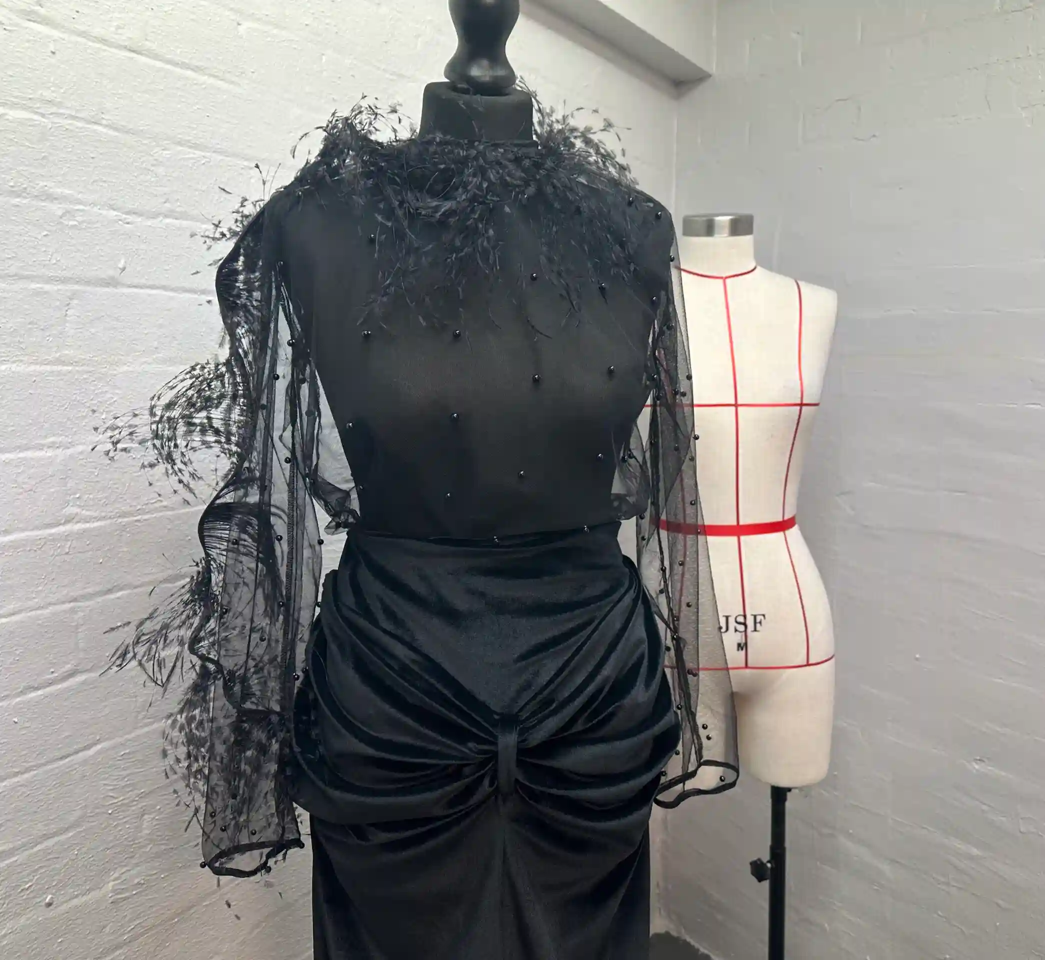 A black mannequin displaying a K&K Couture draped and ruched black satin midi skirt, styled with a black top featuring a feather neckline and sheer beaded tulle sleeves, set against a white painted brick wall in a design atelier