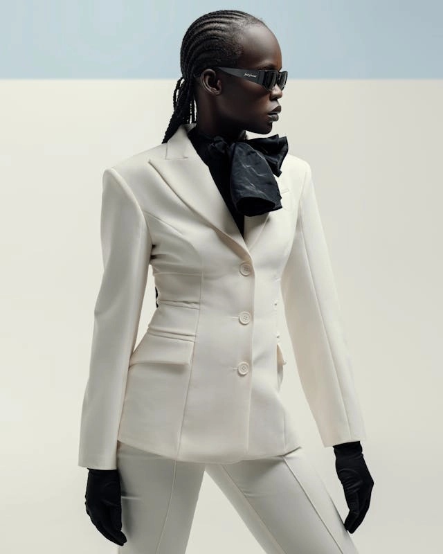 A striking fashion portrait of a woman with braided hair and dark sunglasses, wearing a sharply tailored cream-white K&K Couture blazer. The blazer features strong structured shoulders, peak lapels, and a fitted, sculptural waist with a three-button closure. She styles it with a black neck bow, long black gloves, and matching tailored white trousers against a minimalist, light grey backdrop