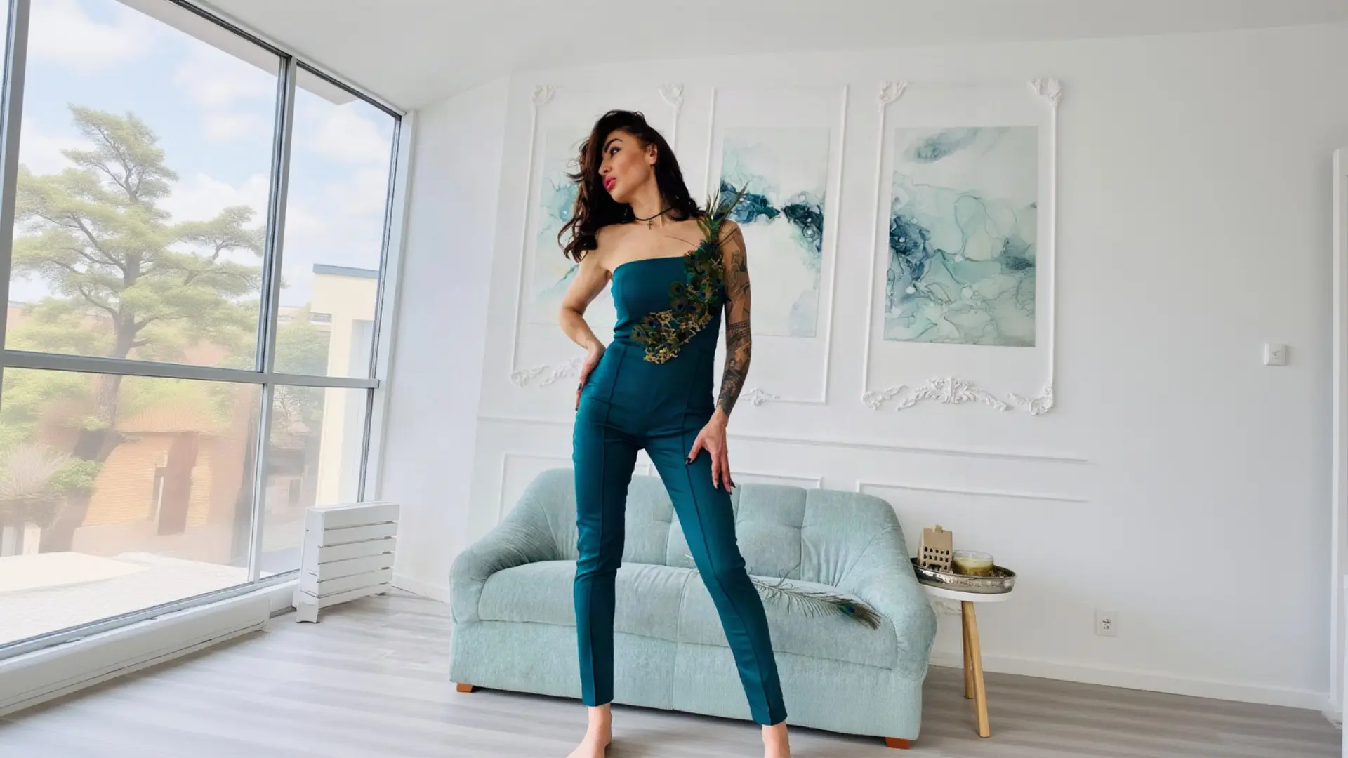 A woman with long dark hair and arm tattoos strikes a powerful pose in a bright, modern living room with a light blue sofa, wearing a deep teal K&K Couture strapless jumpsuit featuring a stunning peacock feather embellishment cascading from the shoulder, while holding a long peacock feather in her hand