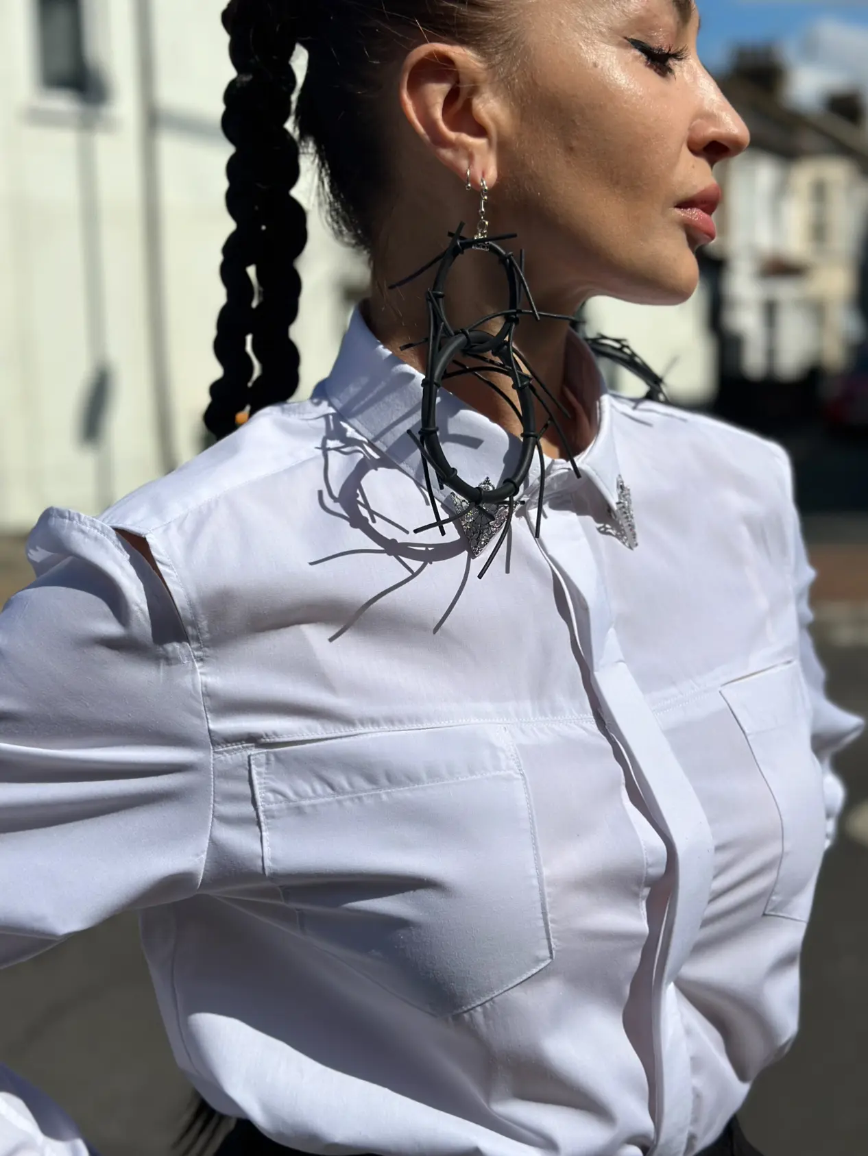 A woman in profile wearing a crisp white K&K Couture button-up shirt with a high mandarin collar, large front patch pockets, and elegant shoulder cutouts. She is accessorized with bold, black wire hoop earrings featuring a silver butterfly charm and stands in bright, natural sunlight against a softly blurred urban background.