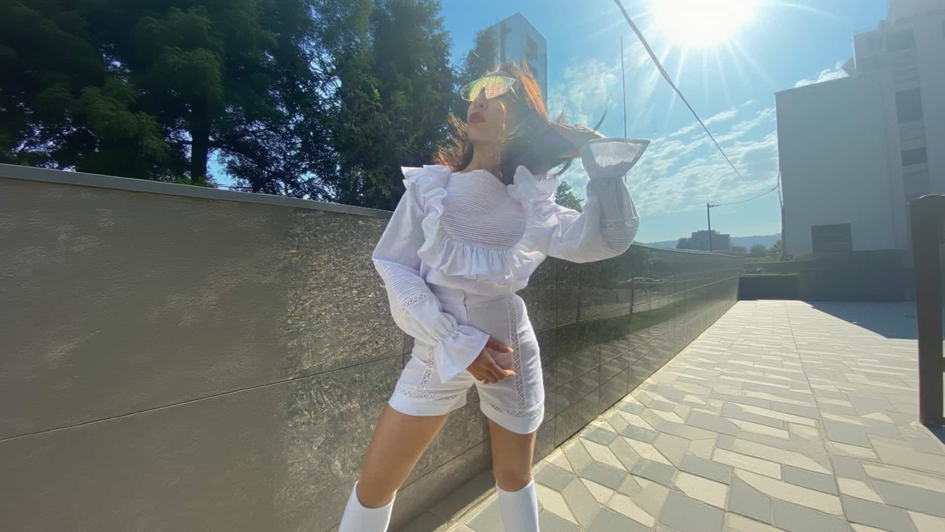 A woman in a white K&K Couture ensemble featuring a smocked, ruffled crop top with sheer bell sleeves, matching white lace-trimmed shorts, white knee-high boots, and iridescent sunglasses, strikes a dynamic, wind-blown pose outdoors against a grey stone wall under a bright, sun-flared sky