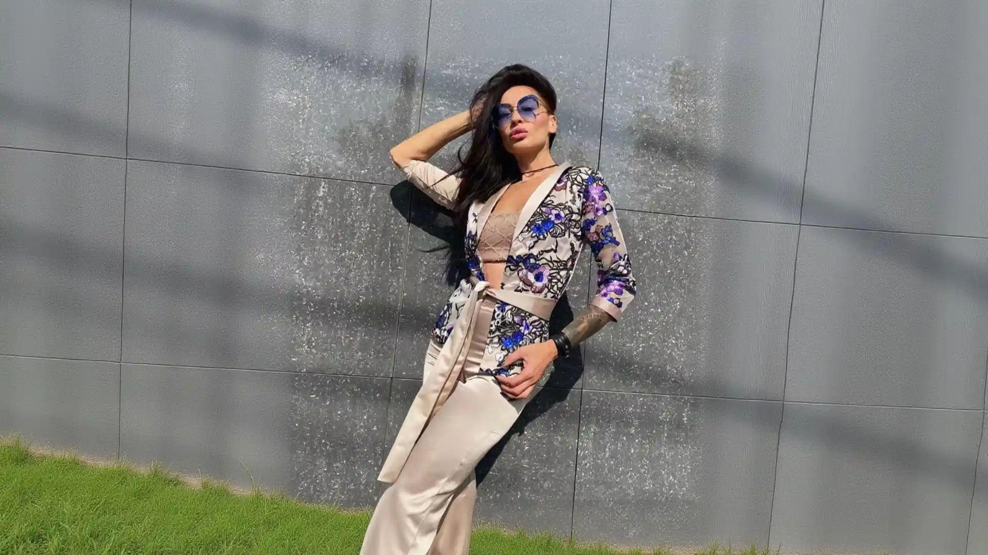 A woman with long dark hair, tattoos, and blue aviator sunglasses leans back against a speckled grey stone wall, wearing a champagne K&K Couture floral blazer and matching satin wide-leg pants over a sheer top, with dappled sunlight and green grass in the background