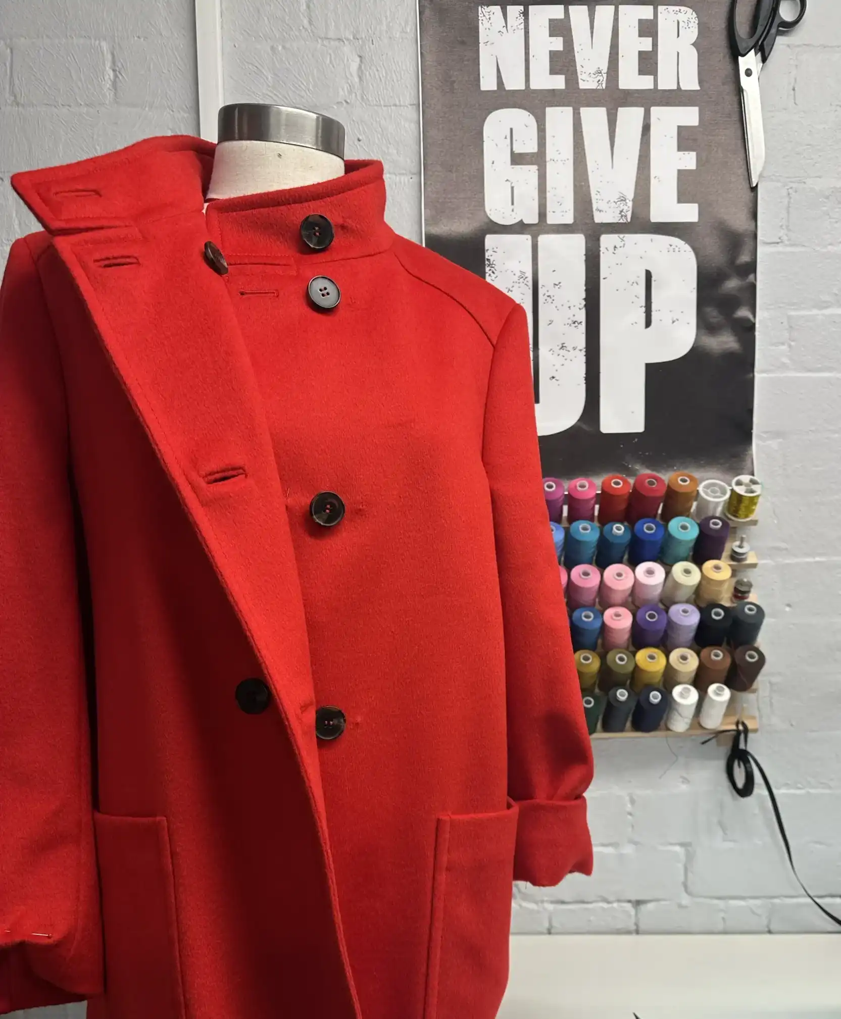 A vibrant red wool coat displayed on a dress form inside the K&K Couture alteration atelier. The double-breasted coat features a high stand collar, large dark buttons, and large patch pockets. A "NEVER GIVE UP" poster, tailoring scissors, and a rack of colourful threads are visible in the background, highlighting the working studio environment.