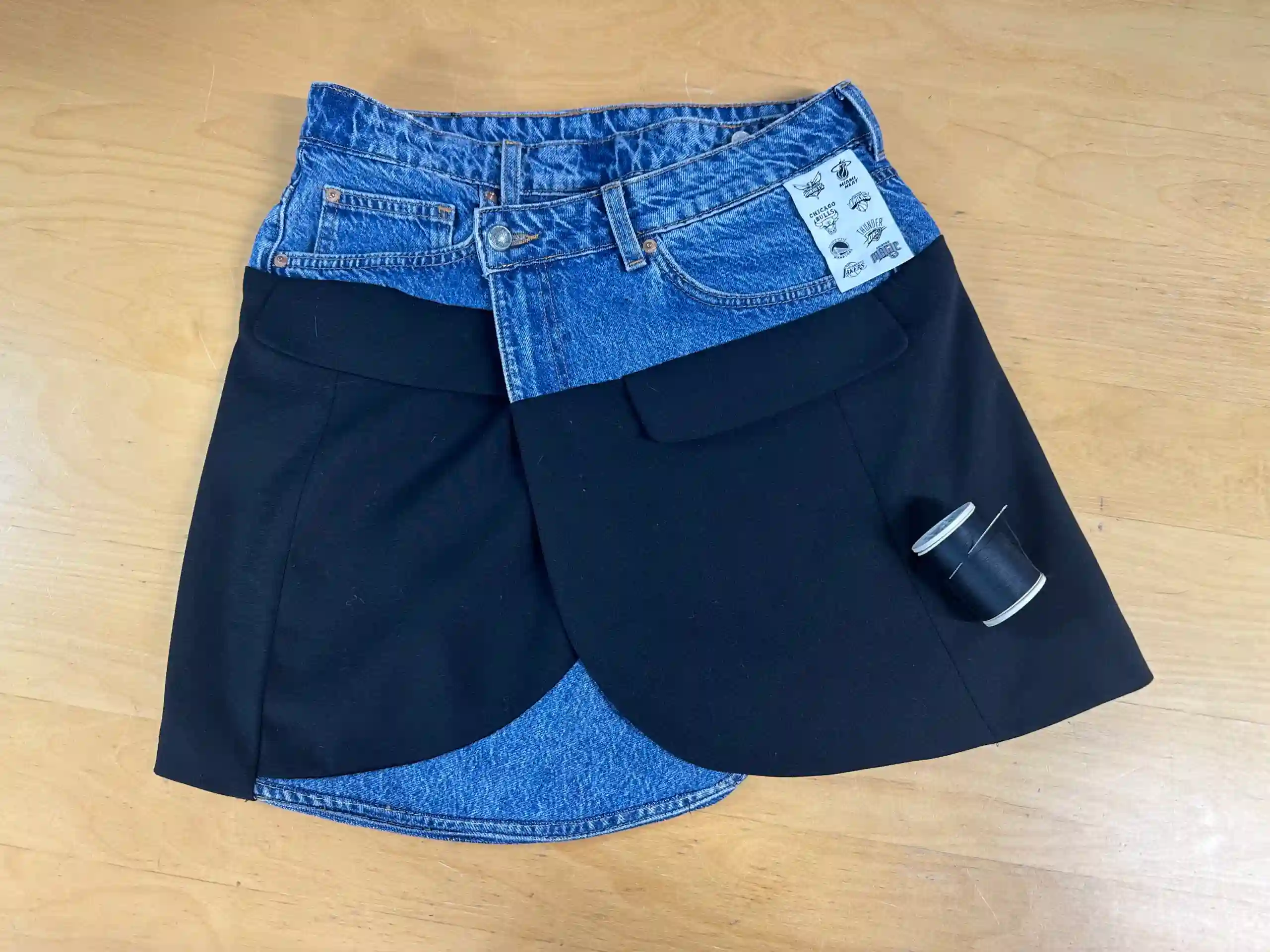 A creative skirt alteration in progress at the K&K Couture atelier, featuring the top half of blue denim jeans with original pockets and a button fly, overlaid with structured black fabric skirt panels featuring large pockets. A spool of black thread rests on the workbench, highlighting the mixed-material design process.