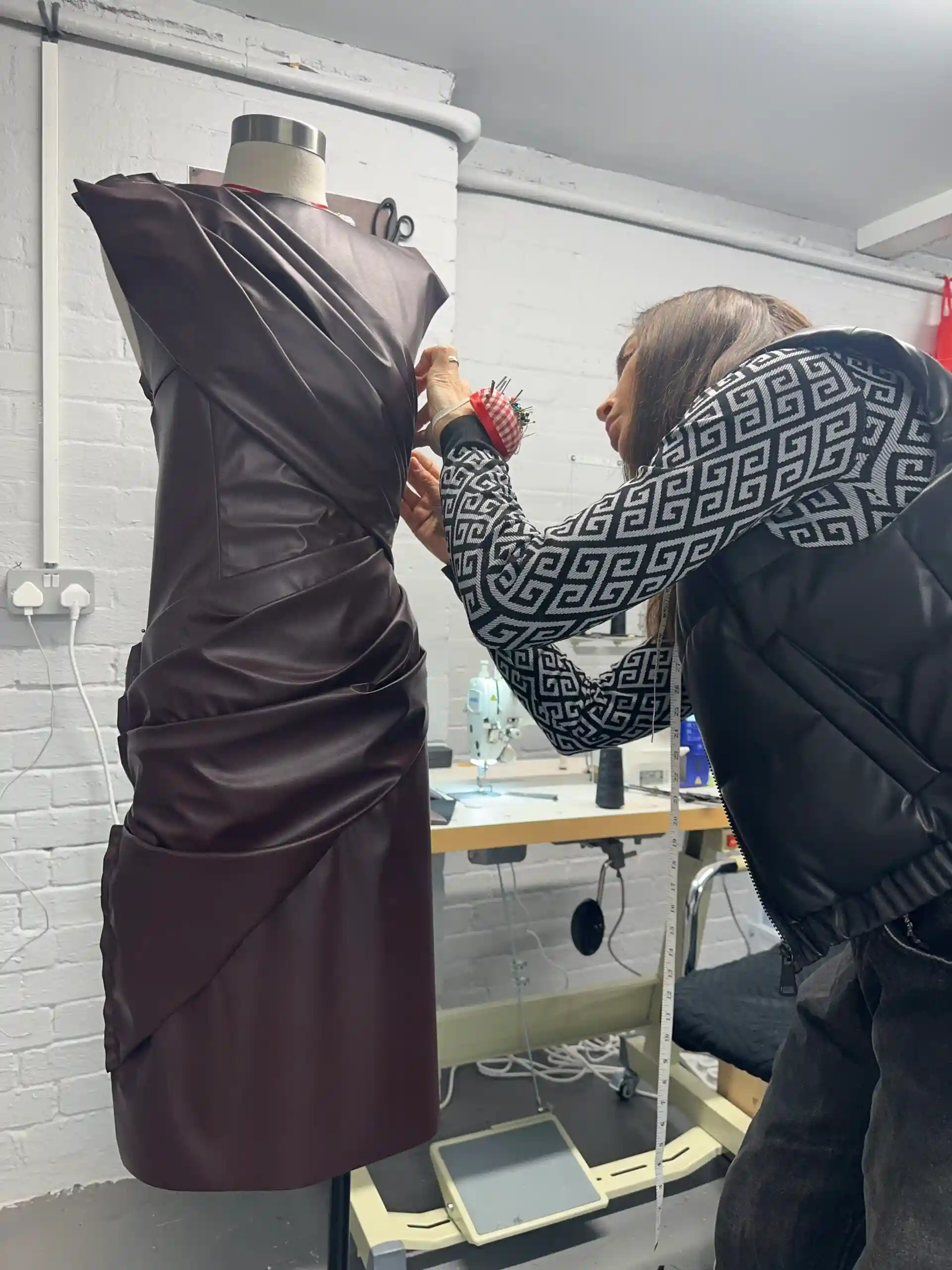 A female tailor at K&K Couture carefully pins a rich, dark brown draped dress on a mannequin in a bright design atelier. The tailor wears a patterned top, a black gilet, and a red wrist pin cushion, with a measuring tape draped around her neck, set against a background featuring an industrial sewing machine and white brick walls.