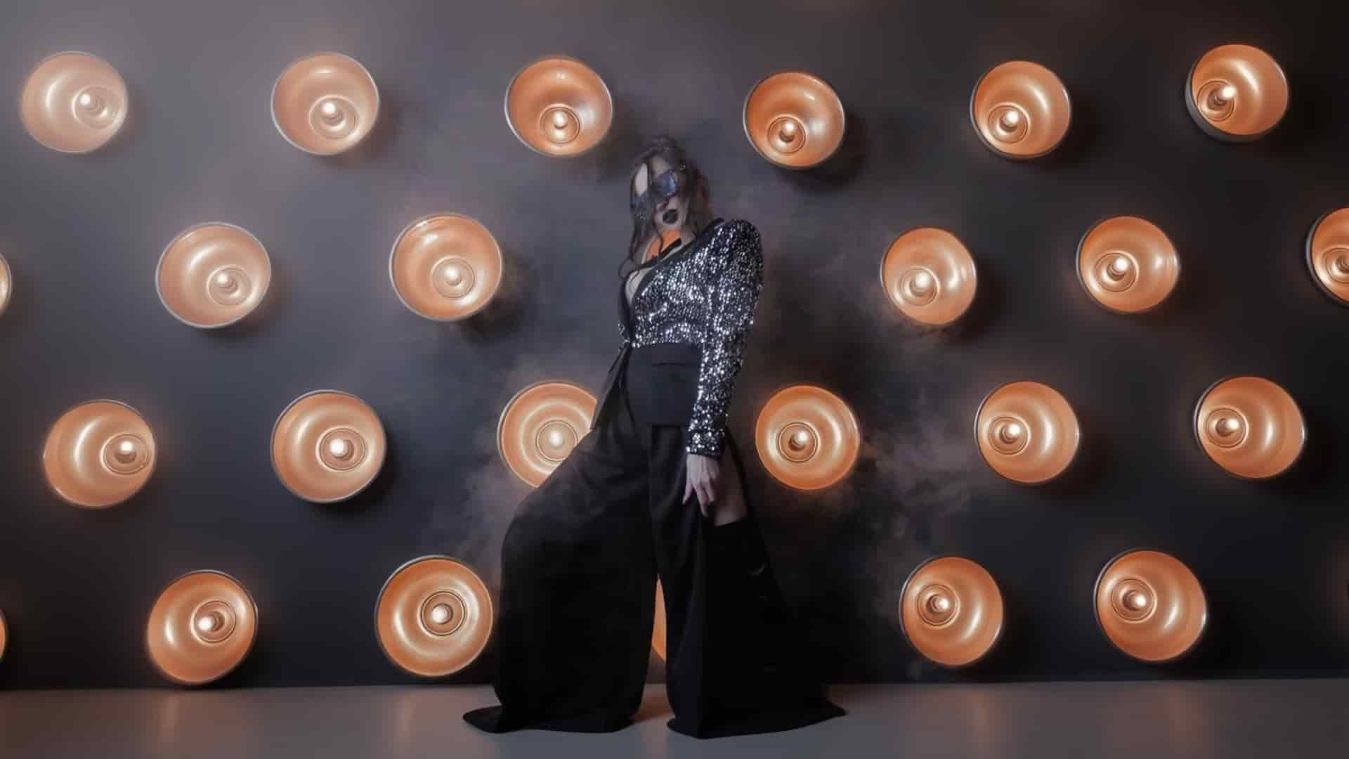 A woman wearing a sparkly long-sleeved K&K Couture black top with a cutout, paired with dramatic flowing wide-leg black trousers and a black lace eye mask, strikes a bold pose in a smoky atmosphere against a dark wall featuring circular copper lights