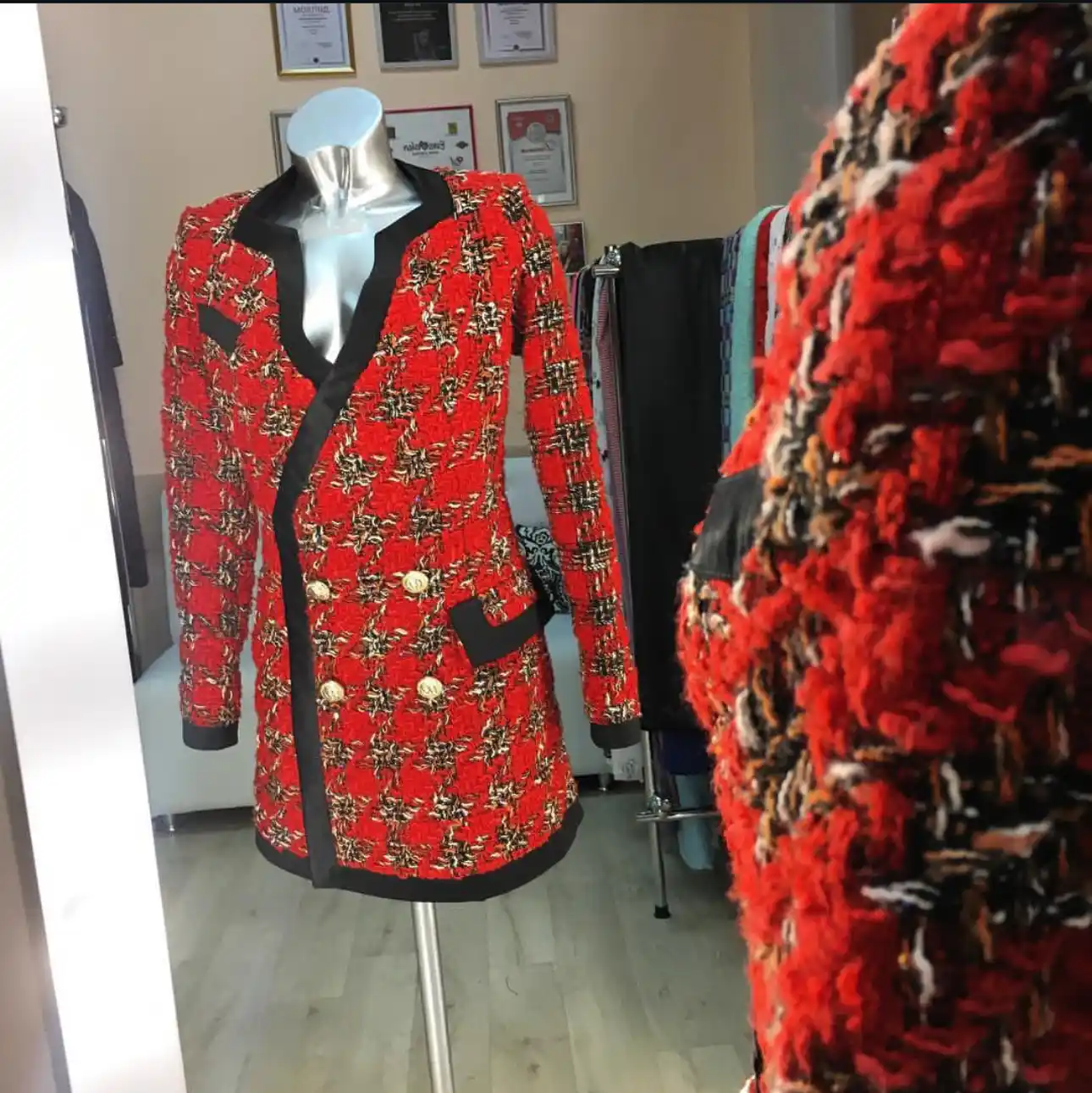 A silver mannequin displaying a striking red, black, and white bouclé tweed K&K Couture coat with black satin lapels and gold buttons, with a close-up of the textured fabric in the foreground, set in a boutique atelier with framed certificates on the wall