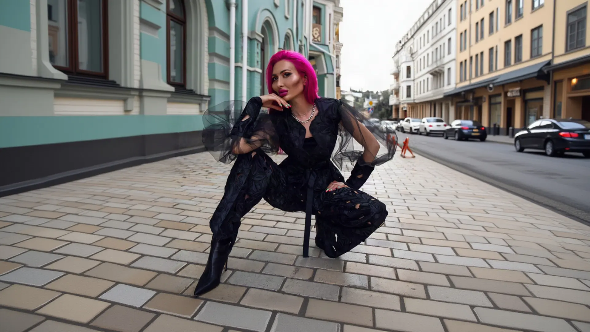 A woman with bright pink hair crouches confidently on a city street, wearing a striking black K&K Couture jumpsuit featuring voluminous sheer tulle sleeves, a textured floral pattern, a waist belt, and knee-high black boots