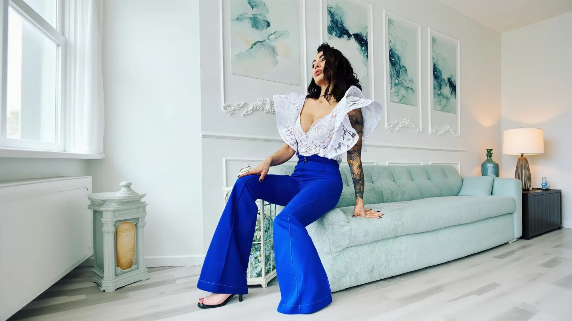 A tattooed woman with dark hair sits confidently on the edge of a light blue sofa in a bright, modern room with abstract art, wearing a white K&K Couture lace top with dramatic ruffle shoulders, vibrant royal blue high-waisted flared trousers, and black strappy heels