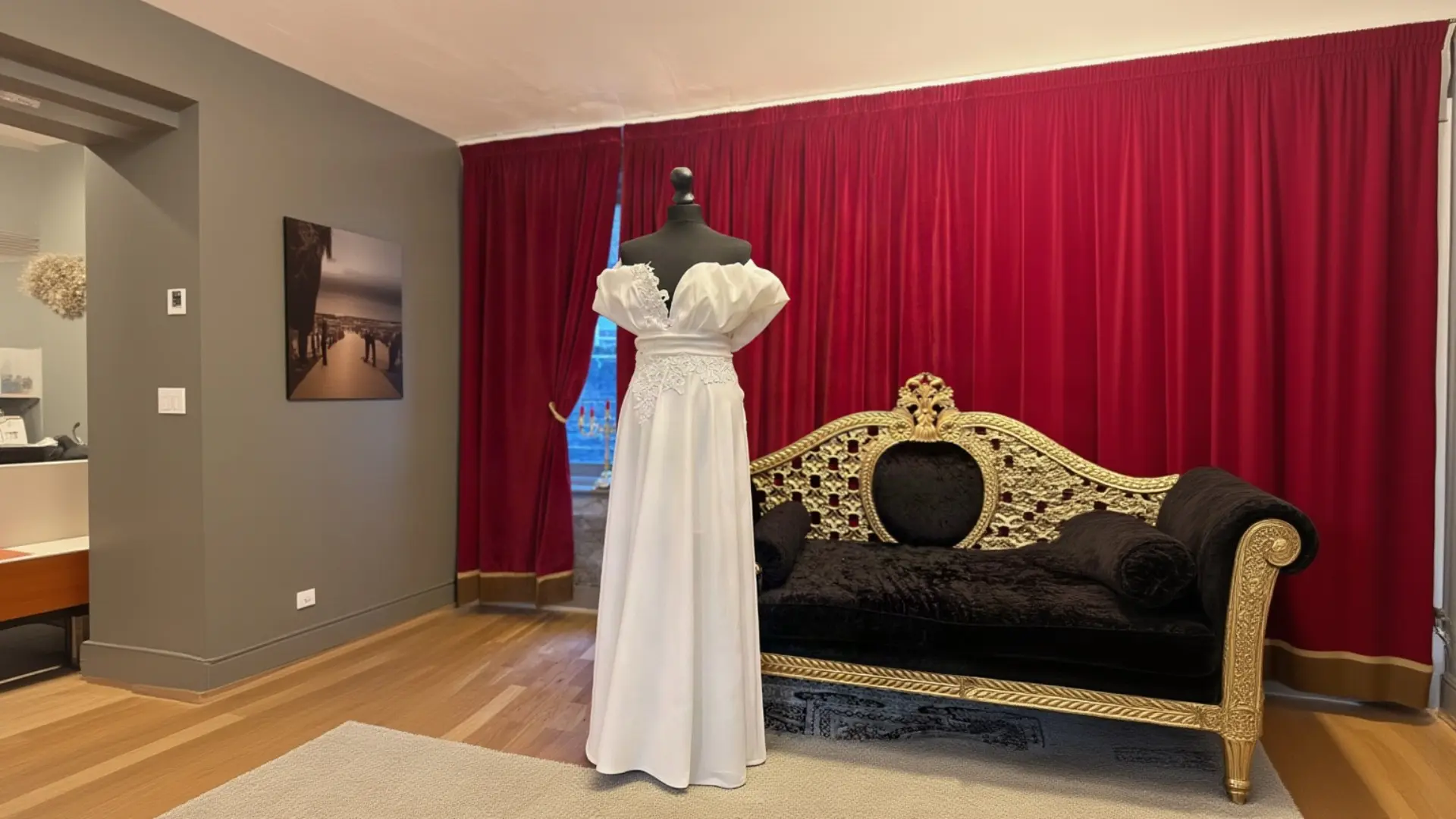 A pristine white off-the-shoulder wedding gown with ruffled sleeves and lace waist detailing displayed on a black dress form, set in an elegant boutique room with deep red velvet curtains and an ornate gold and black vintage settee.