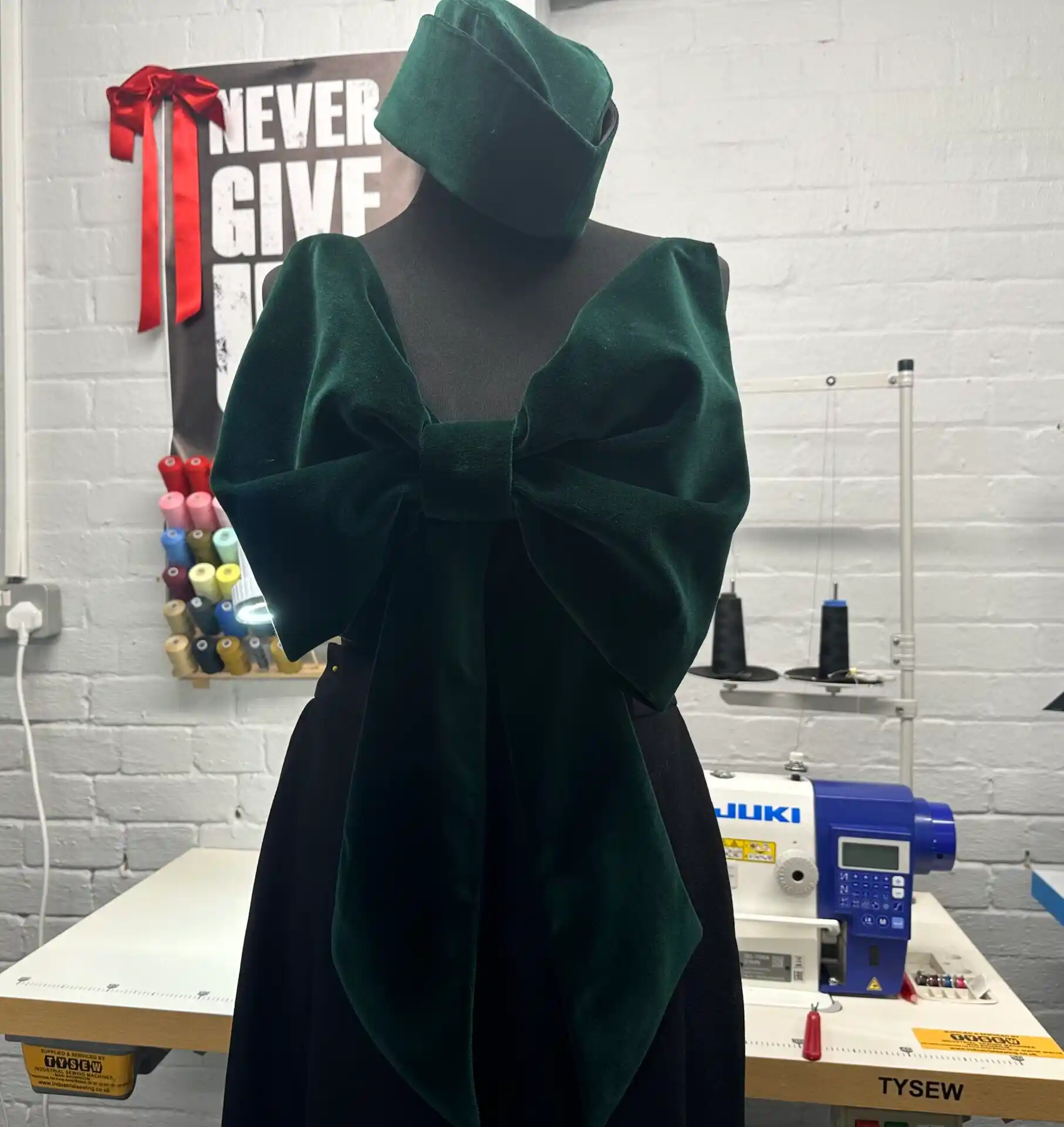 A black mannequin displays a dark emerald green velvet K&K Couture sleeveless blouse, defined by a massive, sculptural bow at the front that creates dramatic shoulder volume without traditional sleeves, paired with a matching velvet headpiece and a dark skirt, set in a bright design atelier.