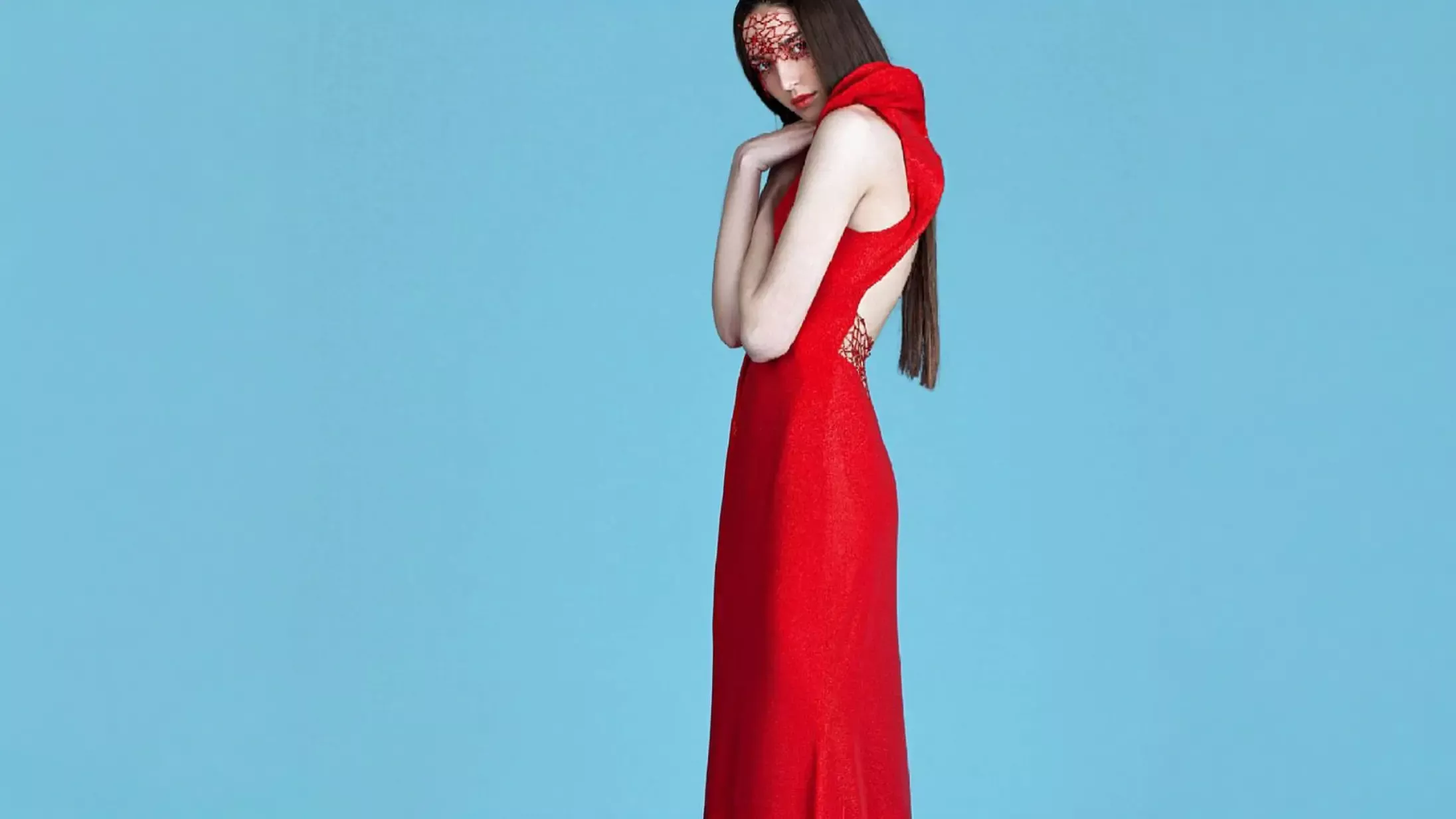 A woman with long straight hair wearing a bright red K&K Couture sleeveless gown with a high structured collar and open back, paired with an intricate red lace eye mask, posing against a solid light blue studio backdrop