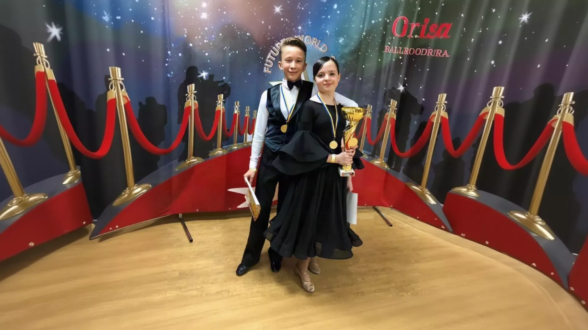 A young boy in a black vest and bow tie and a young girl in a black dress with voluminous sleeves proudly hold a gold trophy and wear gold medals on a wooden dance floor, standing between gold stanchions with red velvet ropes against a starry "Future World" backdrop, styled by K&K Couture