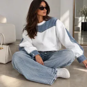 A woman with long dark hair and black sunglasses sits cross-legged on a light floor in a modern, minimalist room. She is wearing a relaxed-fit K&K Couture women's sweatshirt featuring a white body with contrasting blue denim panels on the shoulders, chest yoke, and cuffs, paired with baggy light-wash jeans and white sneakers.