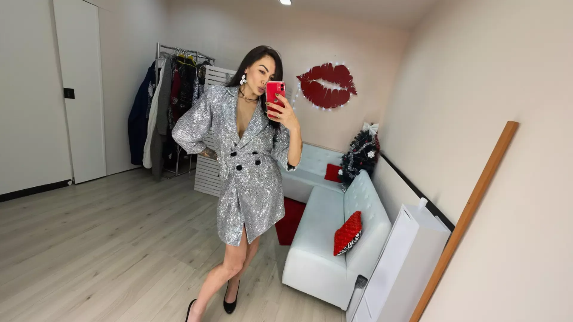A woman taking a mirror selfie with a red phone while wearing a shimmering silver sequin K&K Couture blazer dress with black buttons, black heels, and white statement earrings, in a stylish room with a white sofa, red pillows, red lips wall decor, and a festive Christmas tree