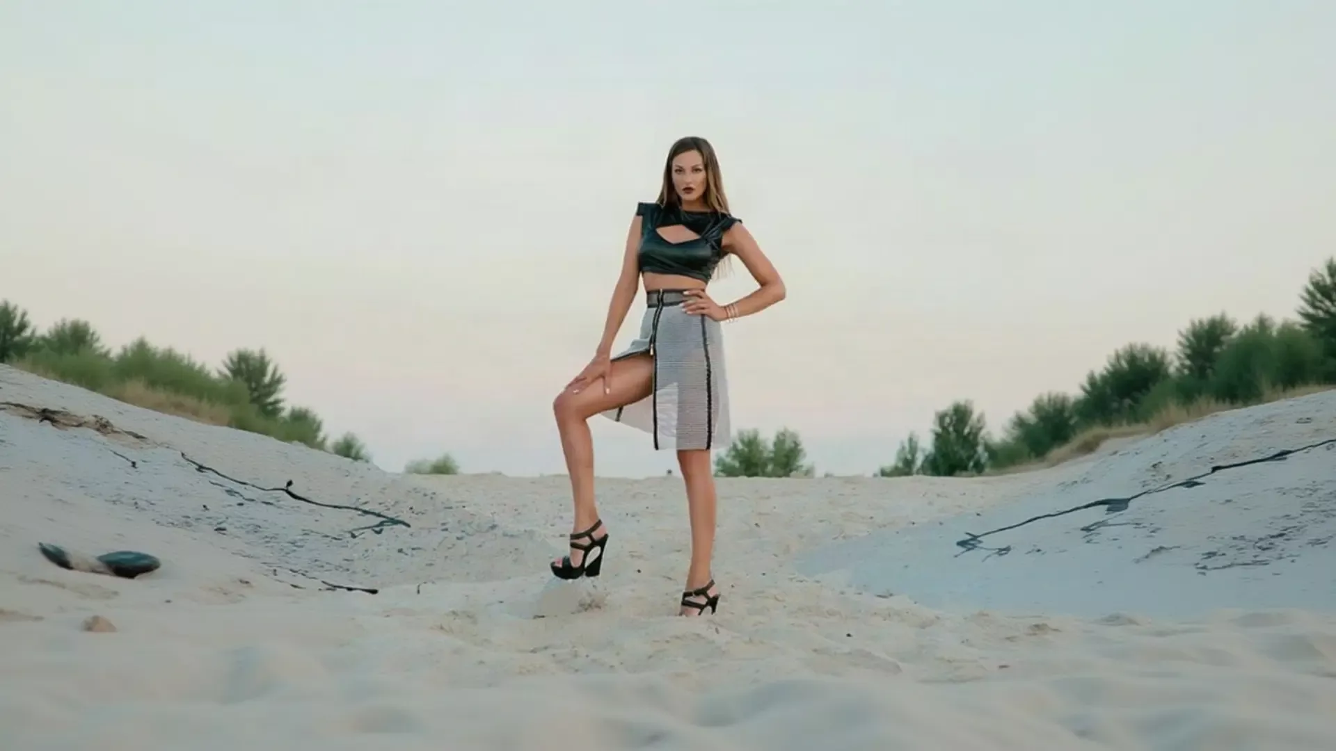 A woman with long hair and bold dark lipstick strikes a confident pose on a sandy landscape at dusk, wearing a black leather-look crop top with a cutout, a high-waisted black and white checkered skirt with a dramatic slit, and strappy black platform heels, showcasing a stunning outfit from K&K Couture