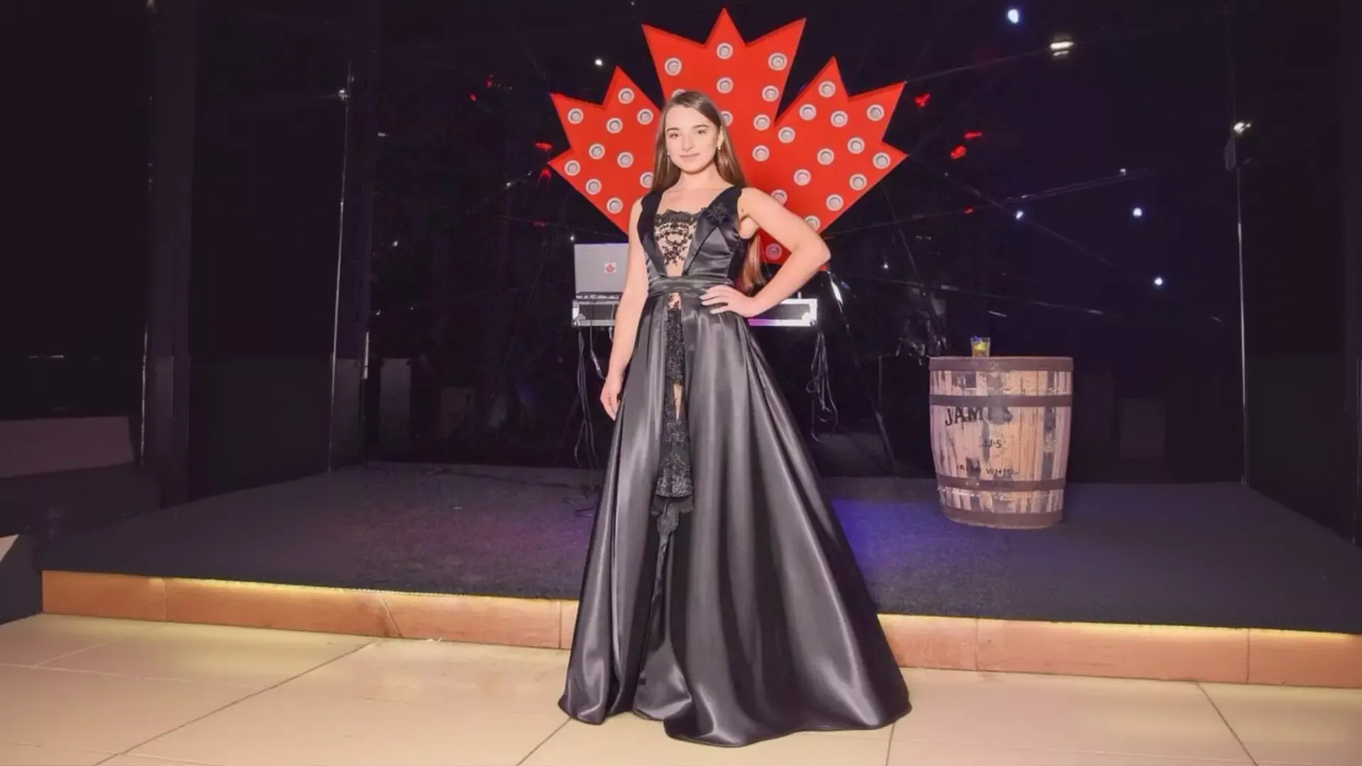 A young woman with long brown hair poses gracefully on an event stage wearing a dark, floor-length K&K Couture satin ballgown with an intricate lace bodice, set against a bright red Canadian maple leaf backdrop