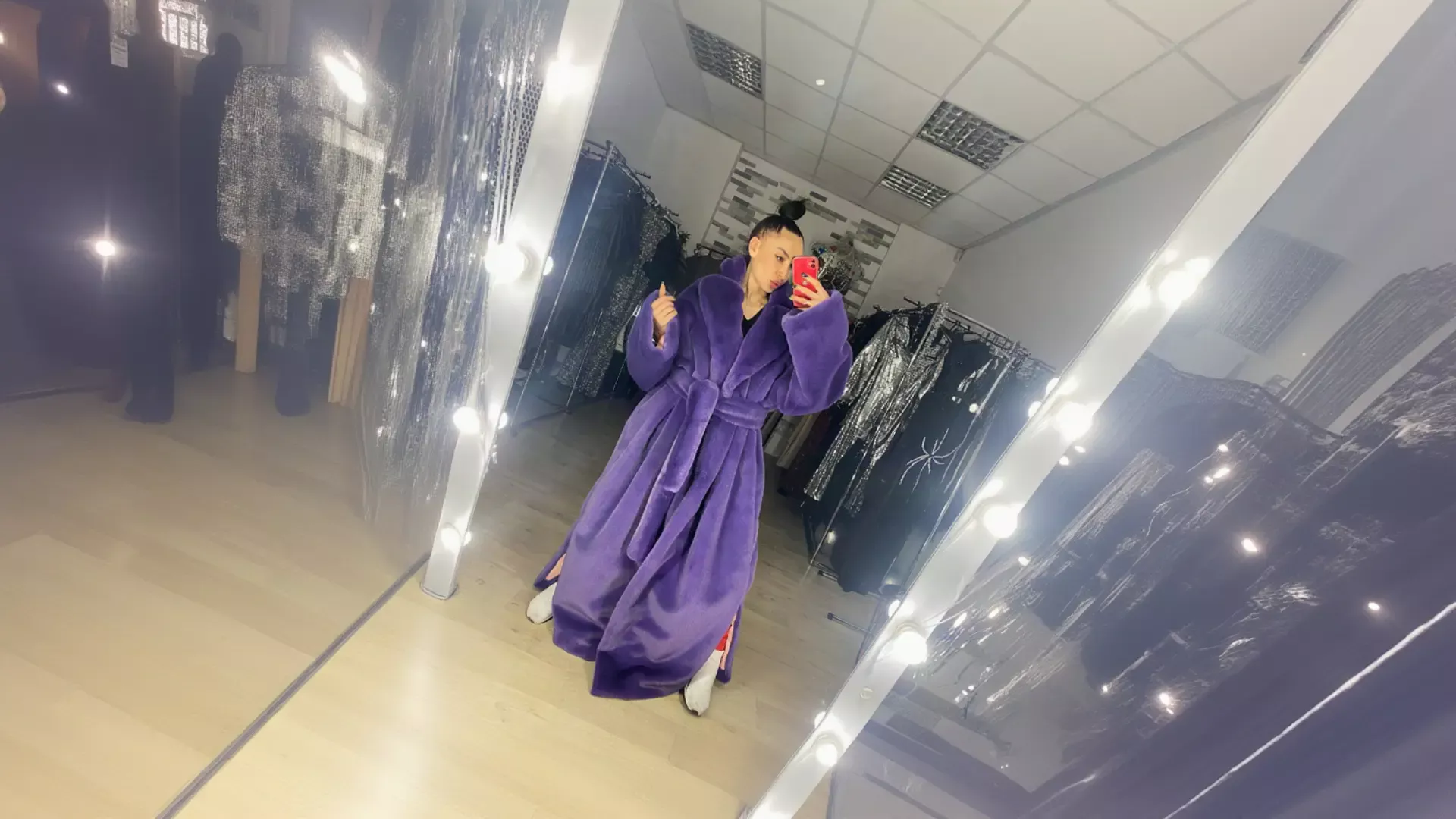 A woman taking a vibrant mirror selfie with a red phone in a boutique, wearing a luxurious, thick purple plush K&K Couture robe with a waist belt and white boots, surrounded by illuminated vanity mirrors and racks of sparkling dresses