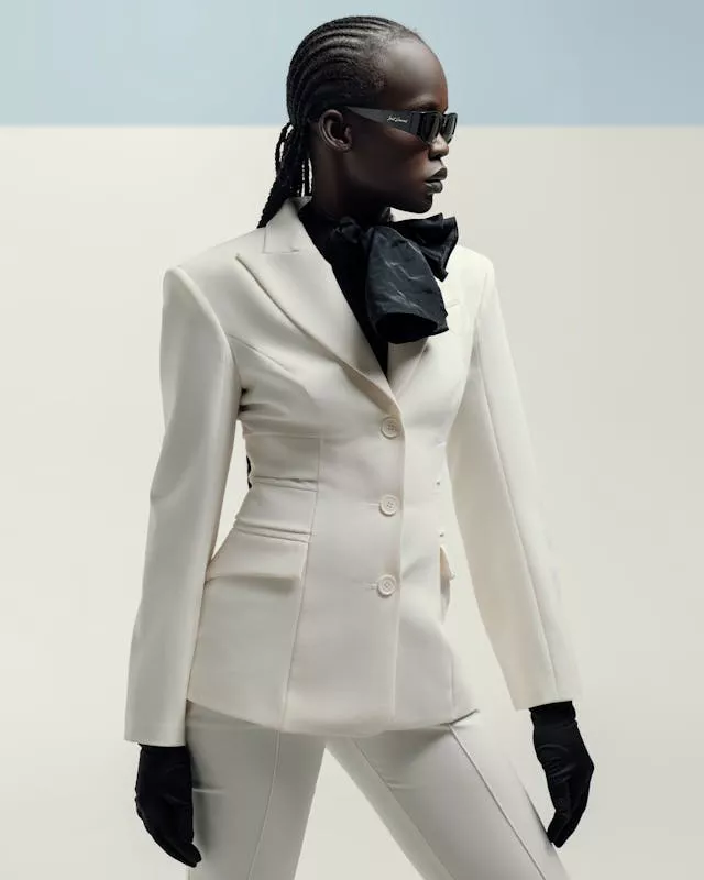 A striking fashion portrait of a woman with braided hair and dark sunglasses, wearing a sharply tailored cream-white K&K Couture blazer. The blazer features strong structured shoulders, peak lapels, and a fitted, sculptural waist with a three-button closure. She styles it with a black neck bow, long black gloves, and matching tailored white trousers against a minimalist, light grey backdrop