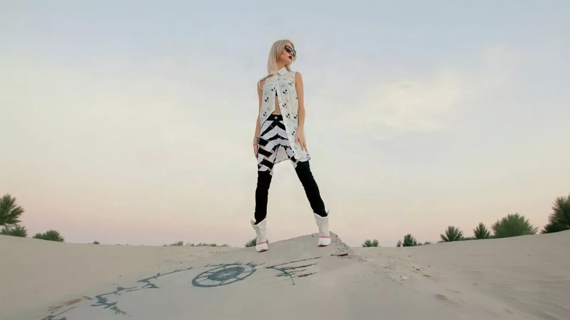 A blonde woman wearing sunglasses and a white geometric K&K Couture vest with black and white chevron pants layered over black leggings, standing on a desert sand dune under a pale twilight sky